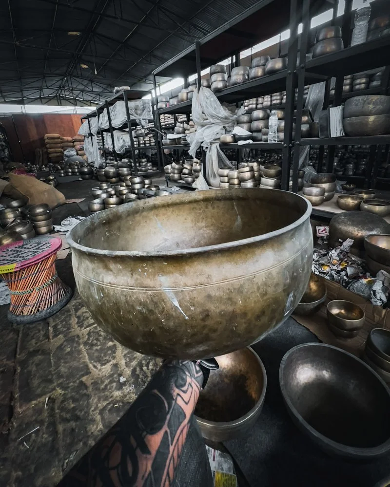 A large metallic singing bowl held by a person with tattooed arms in a warehouse filled with numerous similar bowls on shelves and the floor.