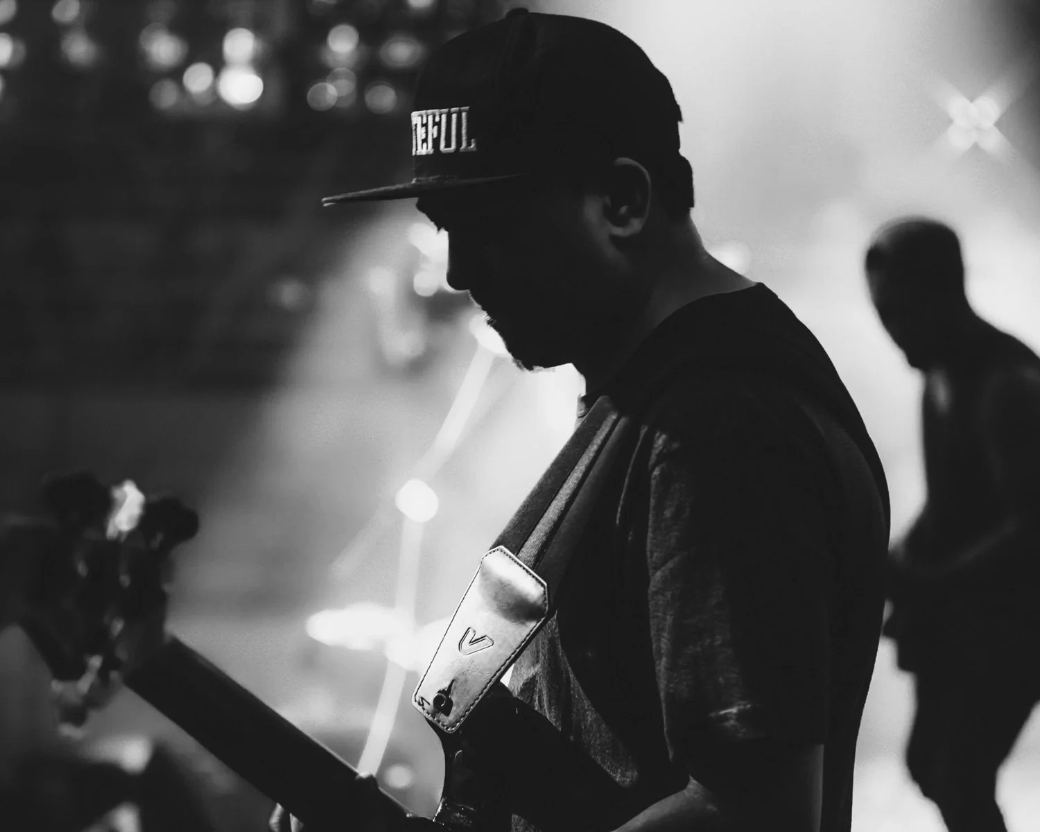 A black and white photo of a man wearing a cap with the word 'HOPFUL' on it, and a backpack, standing next to a motorcycle at night. Another person is visible in the background.