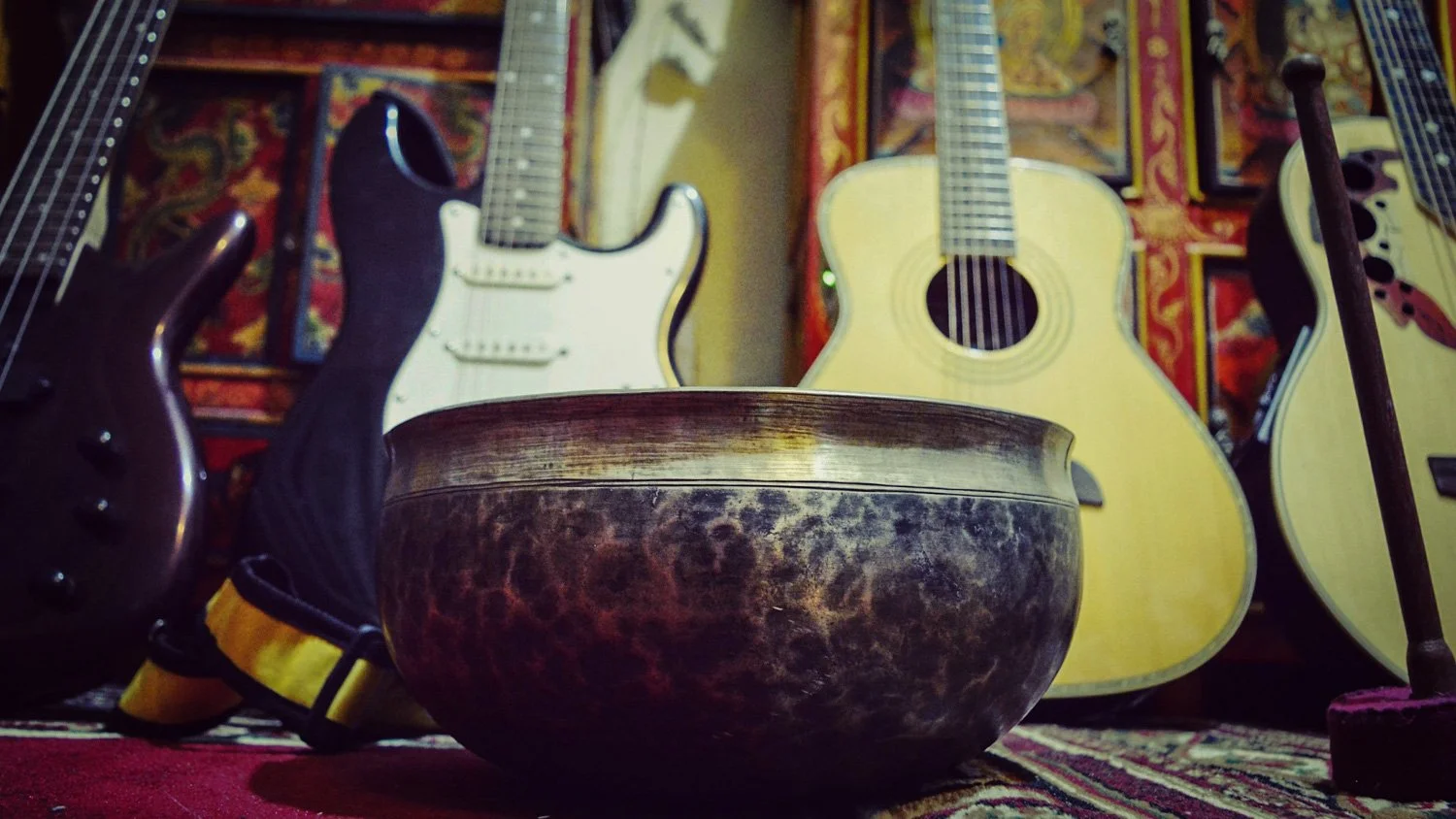 A collection of musical instruments including a large singing bowl, an electric guitar, an acoustic guitar, and a bass guitar, arranged against a colorful, ornate background.
