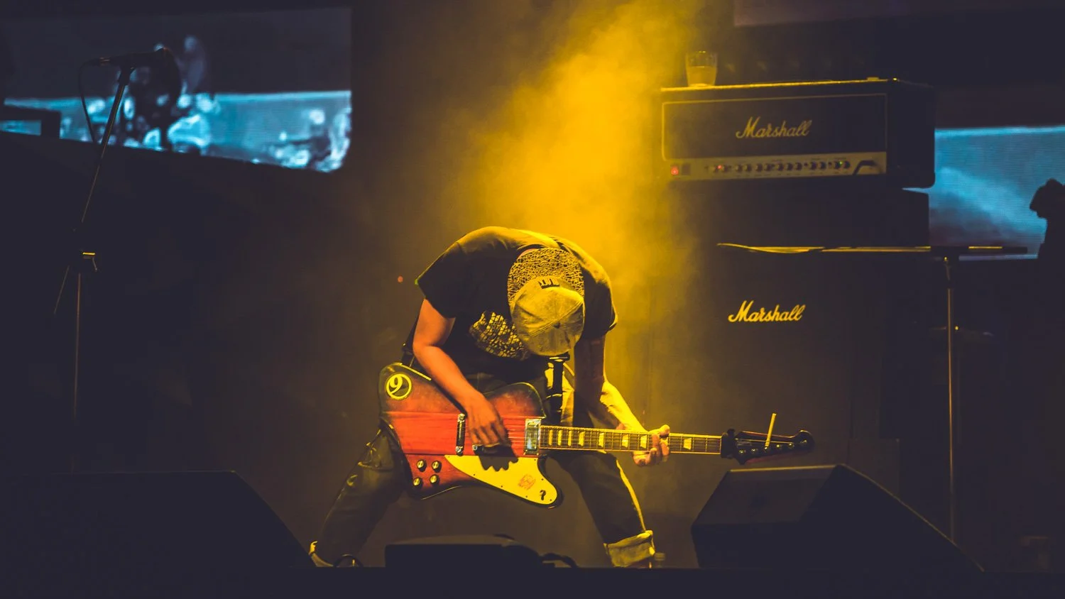 A musician on stage playing an electric guitar, bent forward, with yellow stage lighting and Marshall amplifiers in the background.