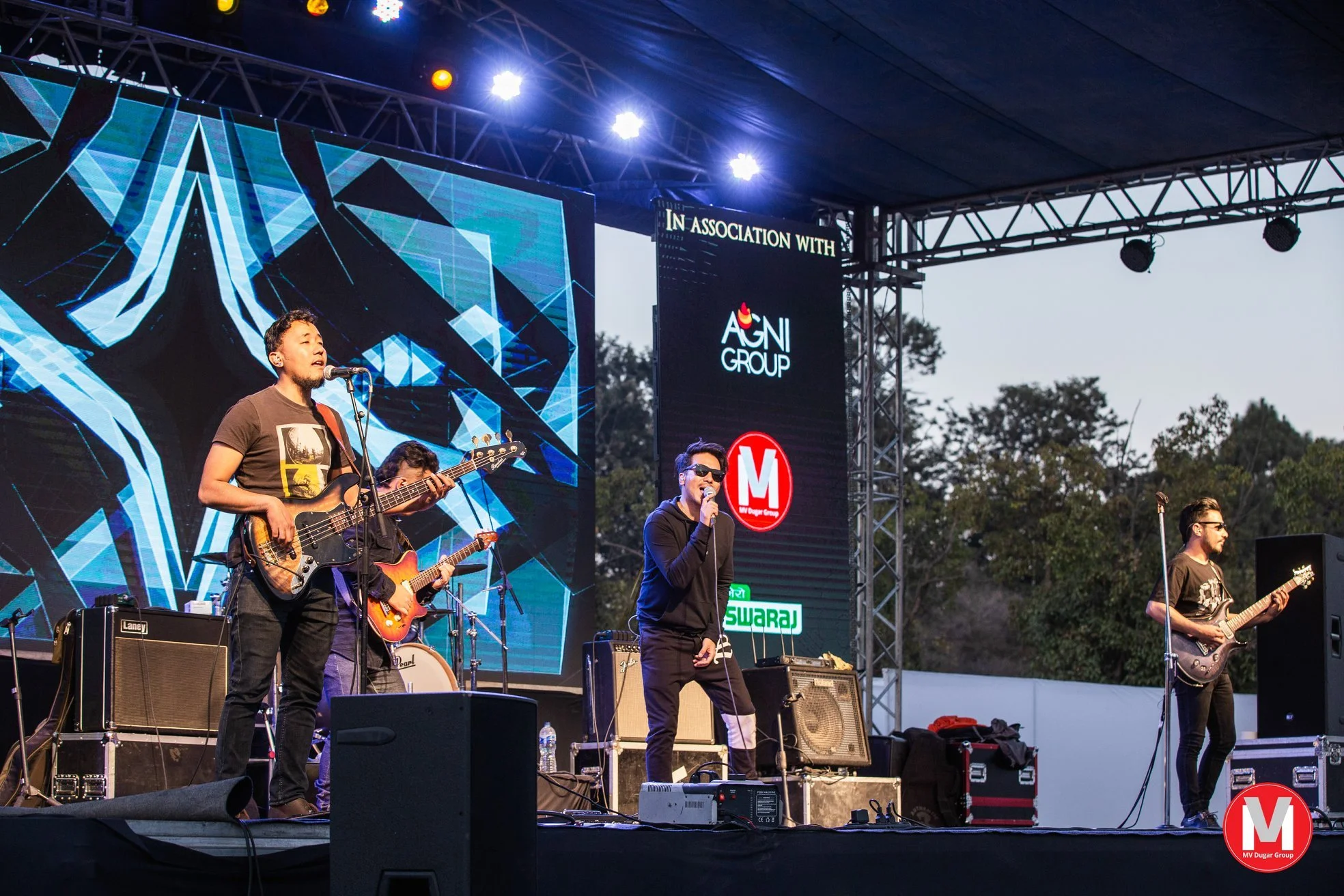 Live outdoor concert with musicians on stage, guitars, drums, and microphones, large digital screen displaying abstract graphic design, stage lighting, trees in the background.