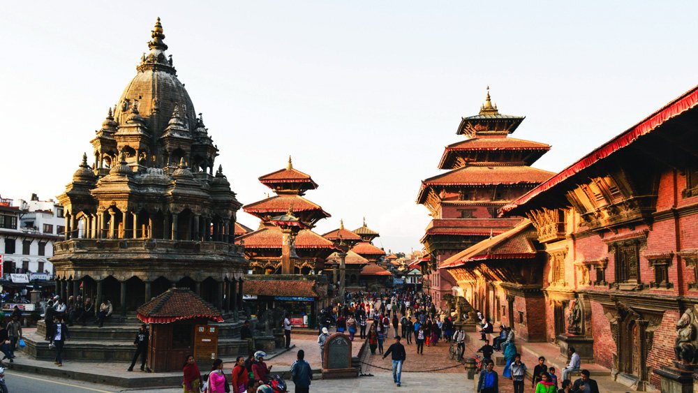 A busy street scene in Kathmandu, Nepal, with traditional pagoda-style temples and buildings, and many people walking around.
