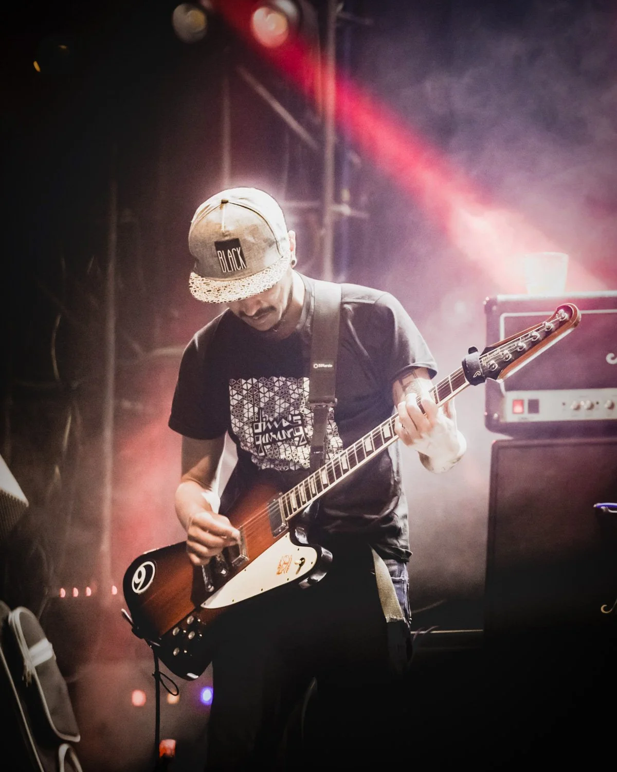 A musician plays an electric guitar on stage with colorful stage lights and amplifiers in the background.