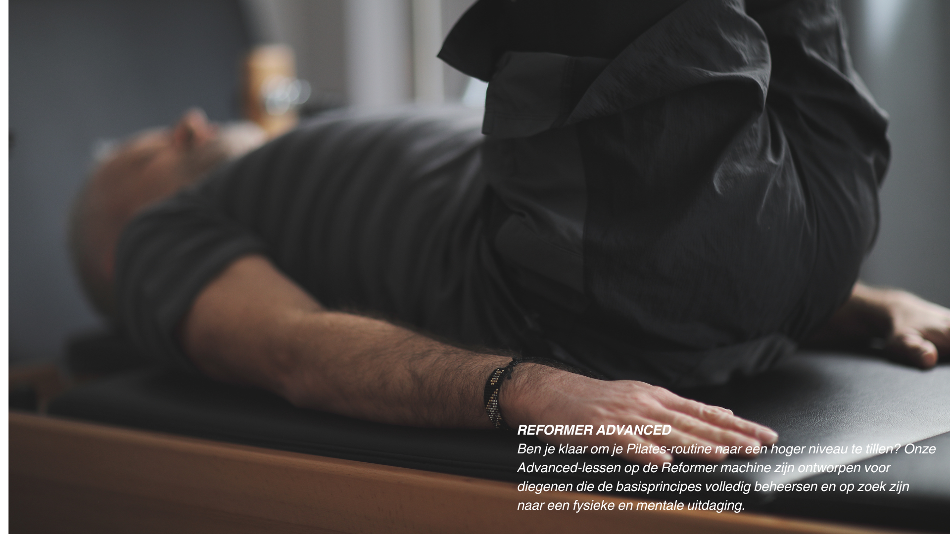 Persoon ligt op een massage tafel, gekleed in een grijze jas, lijkt yoga of stretching te doen, met focus op arm en hand. Raam in achtergrond met natuurlijke lichtinval.