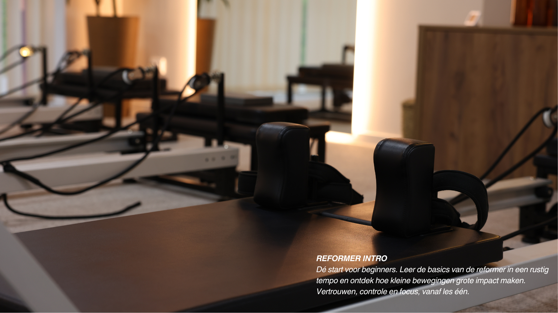 Een rustige zaal met Pilates- of yogaplates, met lichte verlichting en planten op de achtergrond. Op de voorgrond liggen trainingsmachines en kettlebells.