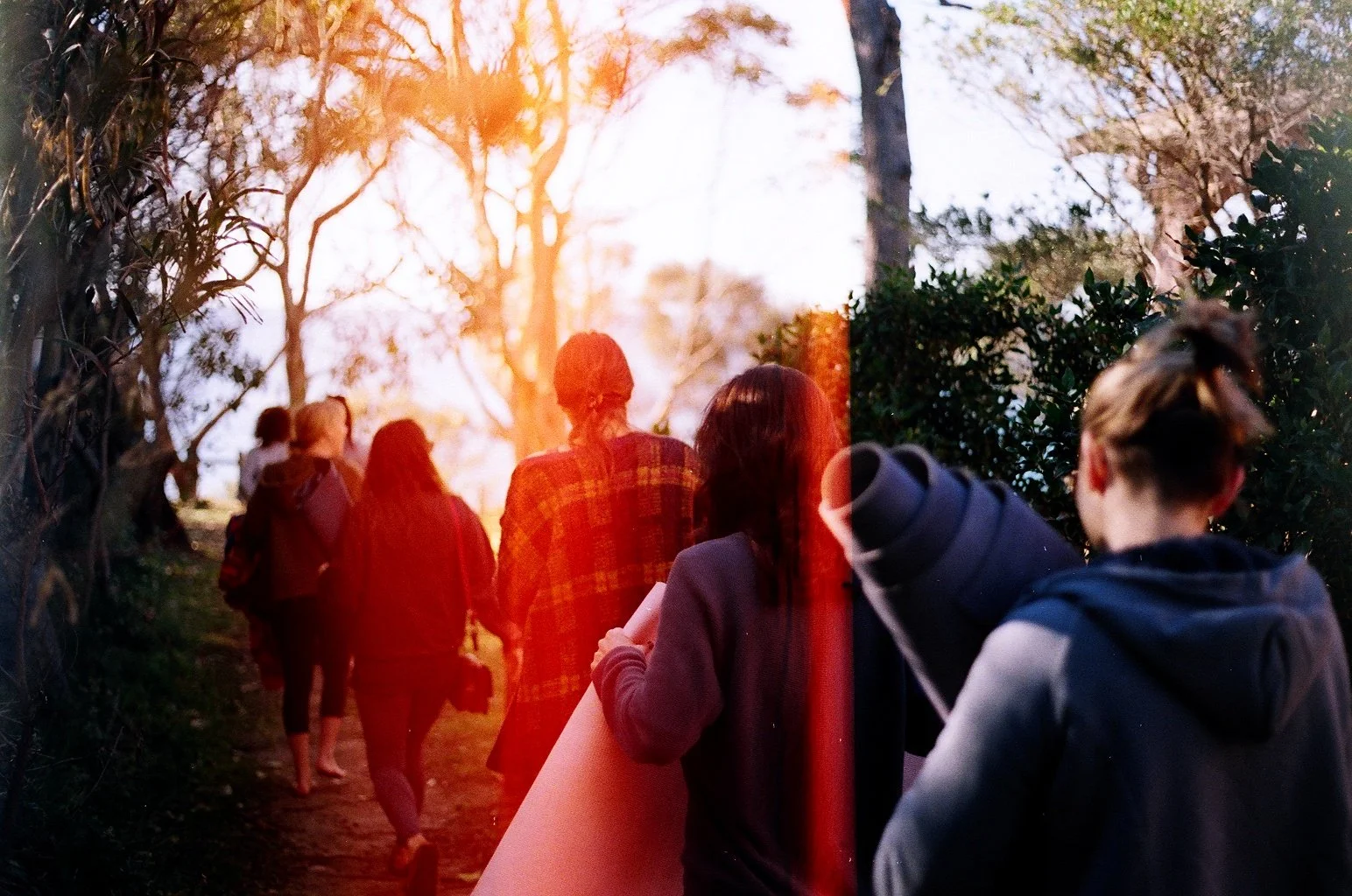 Group of people walking on a path surrounded by trees during sunset, some carrying backpacks and a rolled-up poster.