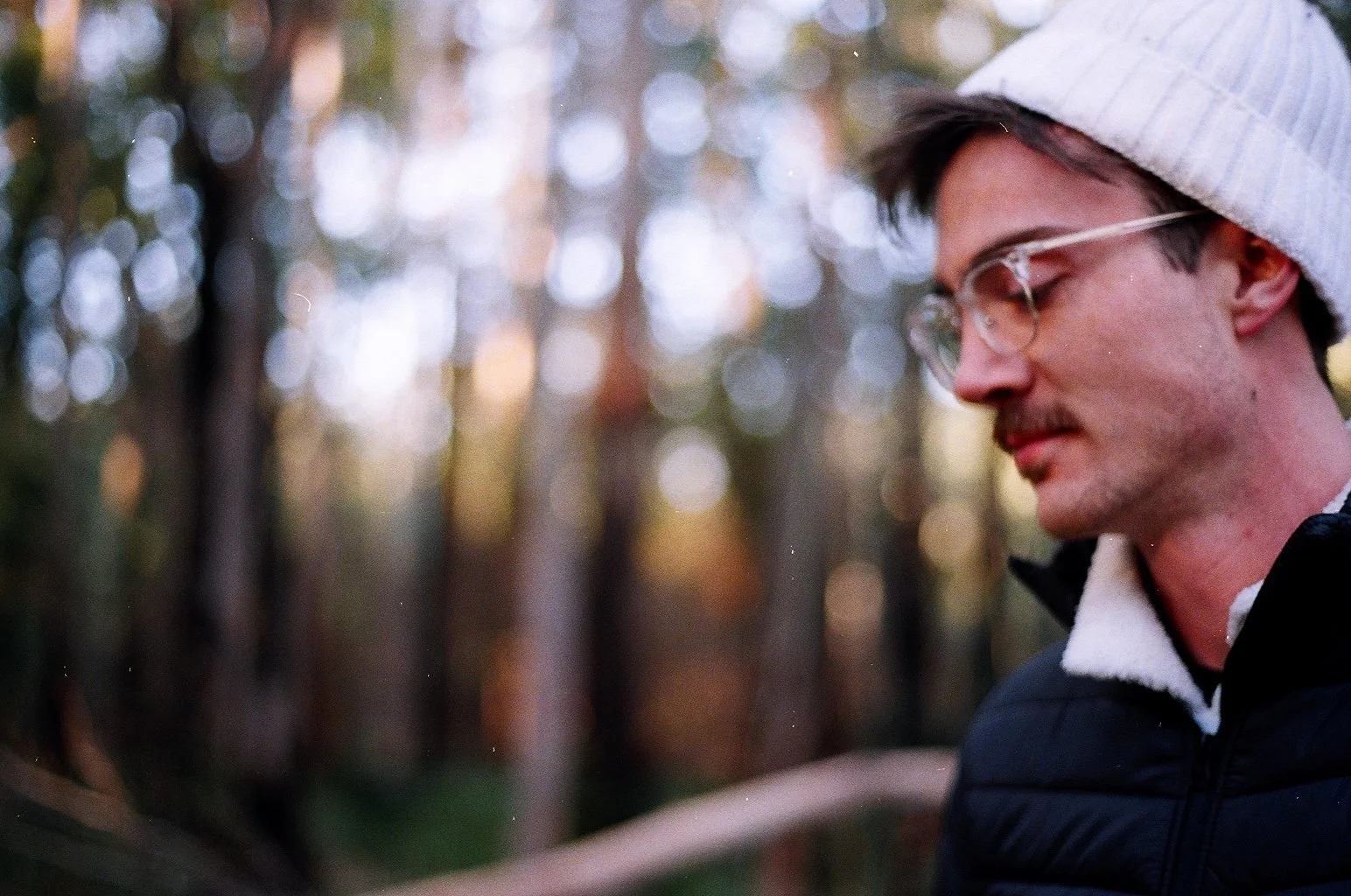 A young man with glasses and a white beanie looking down in a forest background with bokeh light effects.