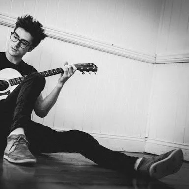 Hedley Nelson sitting on the floor playing an acoustic guitar in a room with plain walls