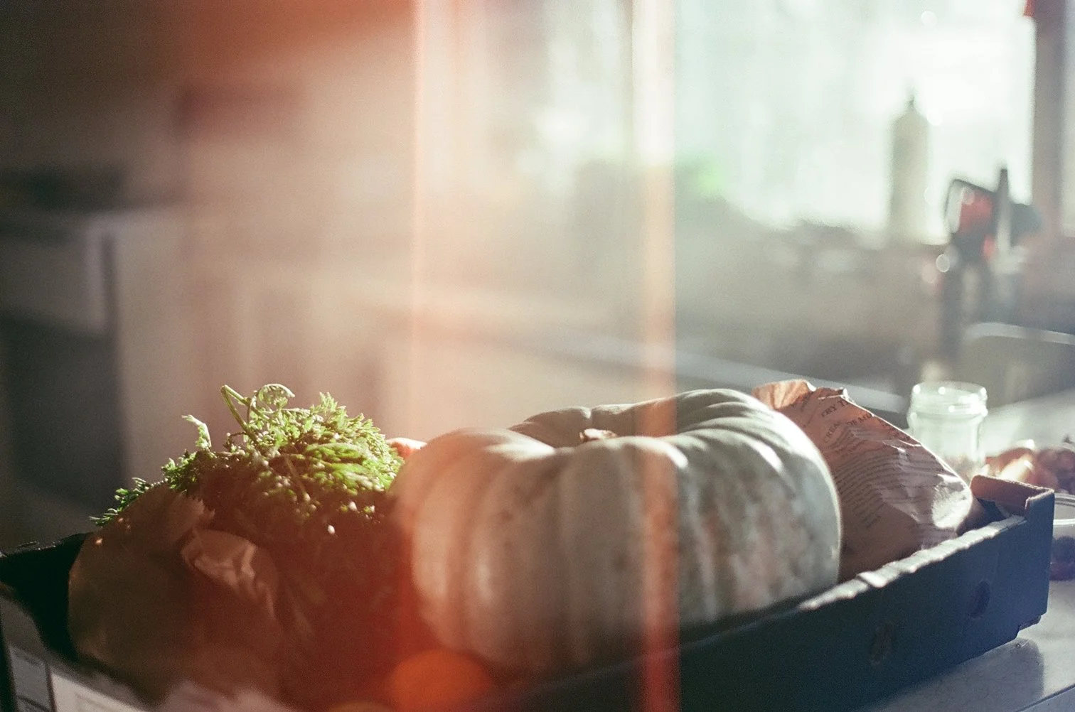 A tray of raw vegetables and bread wrapped in paper, placed on a countertop in front of a window with sunlight streaming in.