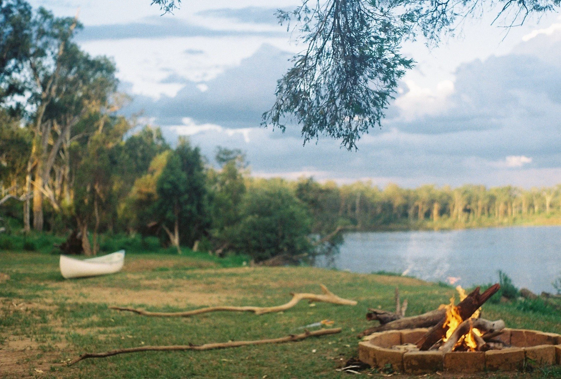 A lakeside scene during sunset with trees lining the shore, a small boat on the grass, and a campfire burning in a circular fire pit in the foreground.