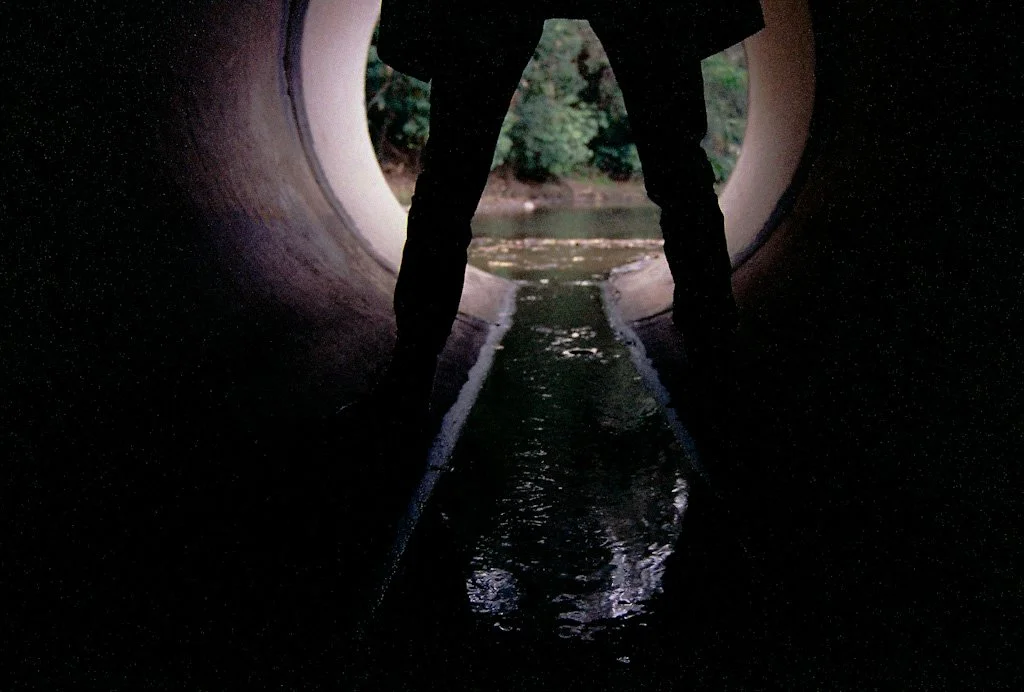 Person standing inside a large concrete pipe with a view of water and trees outside, taken from inside the pipe.