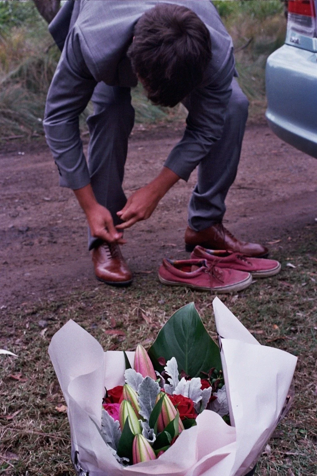 A man is bending down outdoors to tie his brown leather shoes next to a bouquet of flowers on the ground, with a car partially visible on the right side of the image.