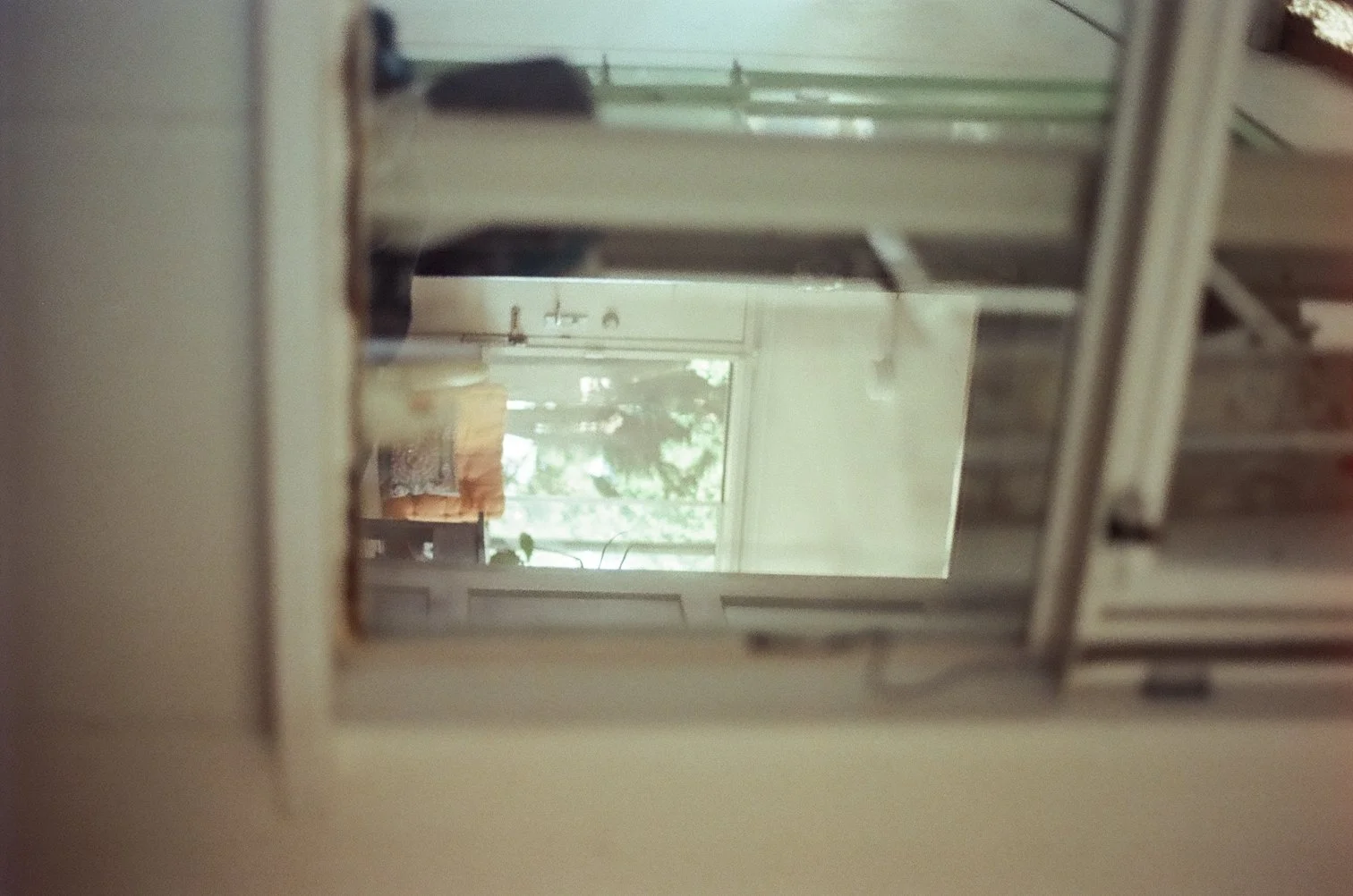 Blurred interior view of a room seen through a mirror, showing a window with outdoor greenery, a chair with a orange cushion, and a piece of furniture with a lamp.