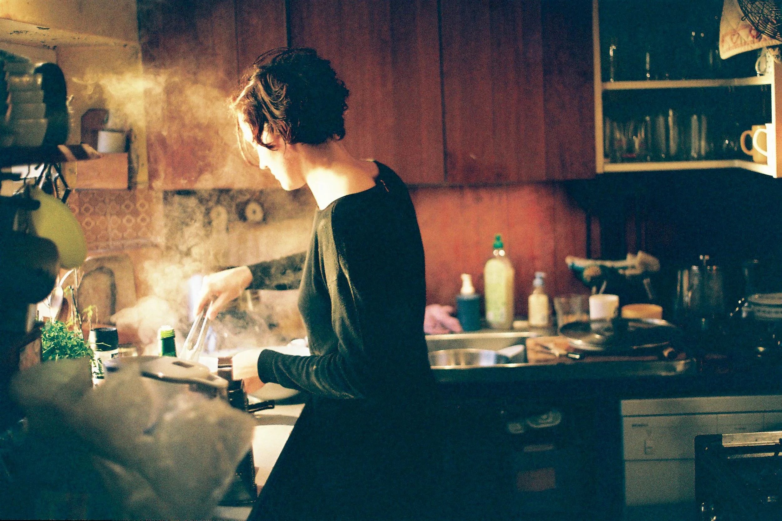 A woman with short, curly hair cooking in a cozy, dimly lit kitchen. She is focused on her task, surrounded by various kitchen items and plants.