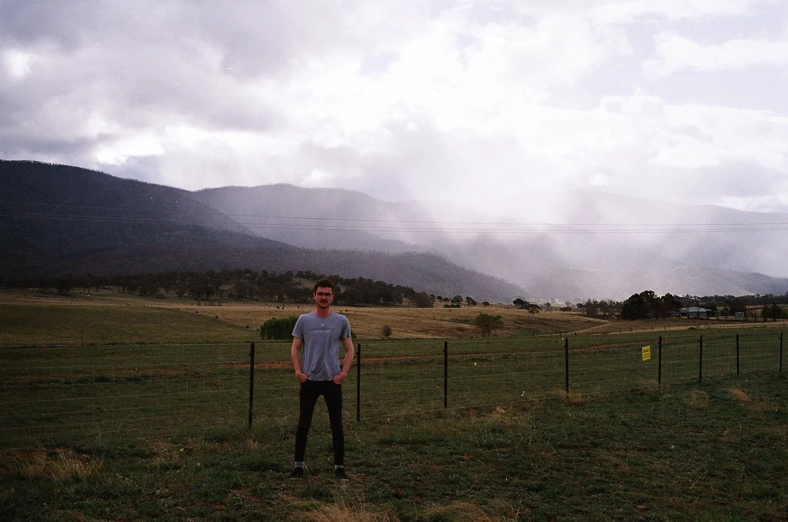 A man standing in front of a fence in a rural landscape with mountains in the background and cloudy sky above.