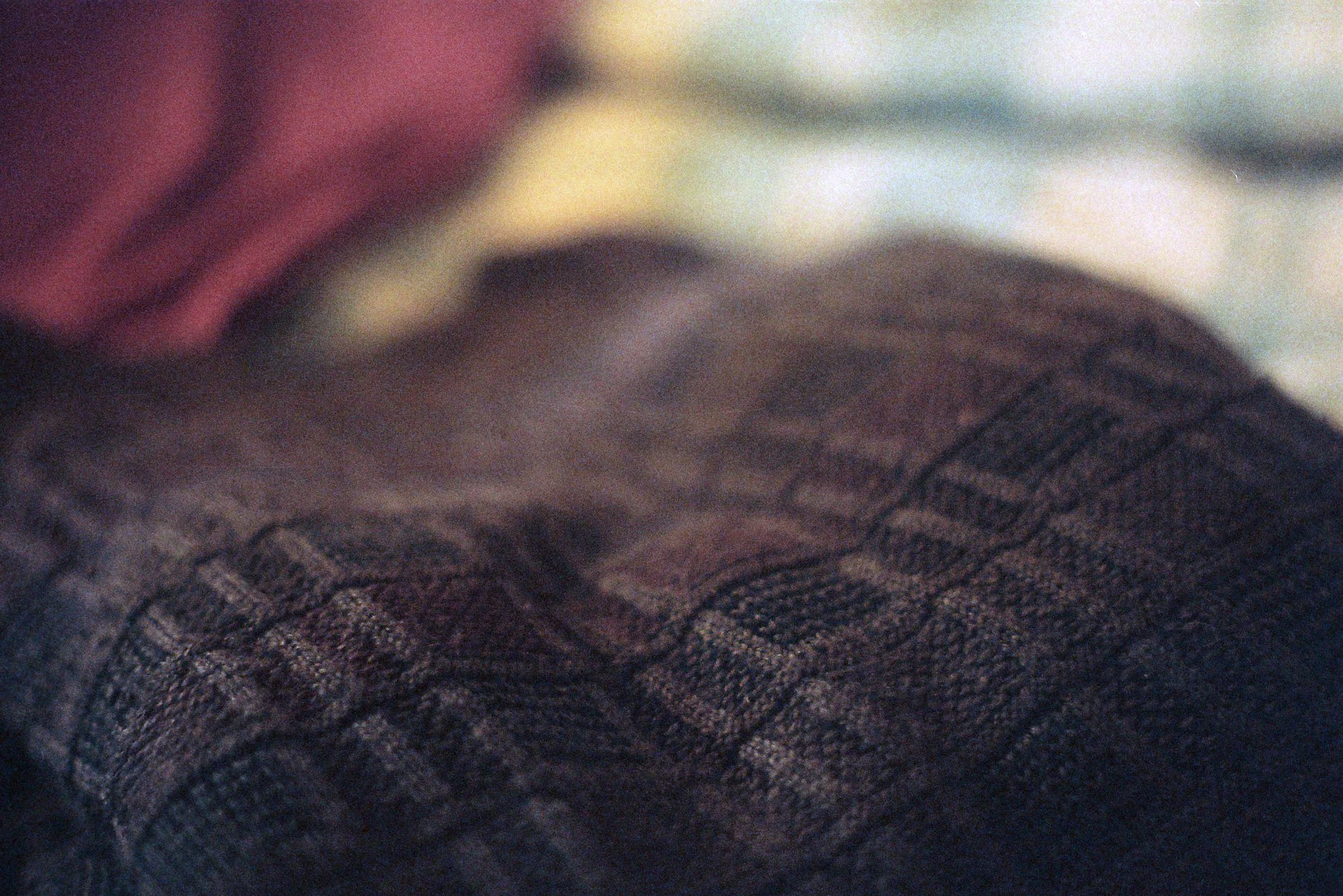 Close-up of dark-colored fabric with textured, patterned surface and a blurred background with red, beige, and blue tones.