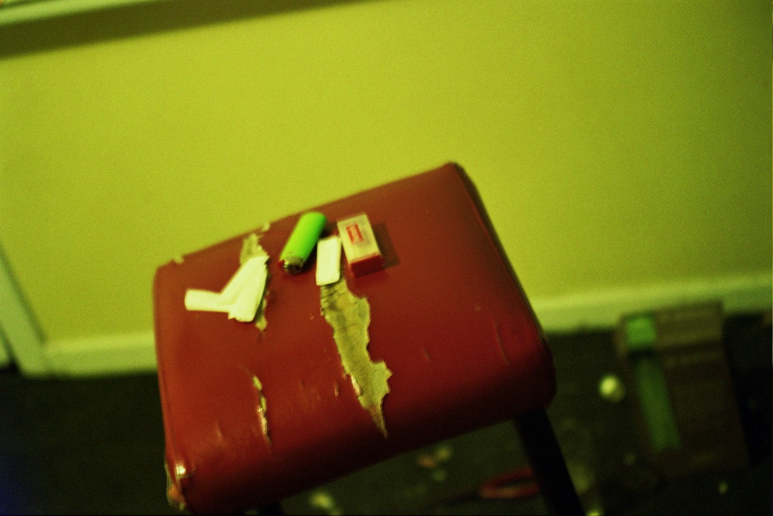 A red stool with peeling paint, holding a torn cigarette pack, a lighter, a green cigarette, and a torn piece of paper, in front of a yellow wall.
