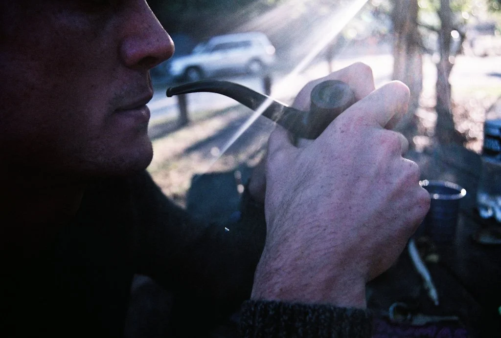 Close-up of a person's face and hand holding a pipe with sunlight shining through, outdoors with parked cars and trees in the background.