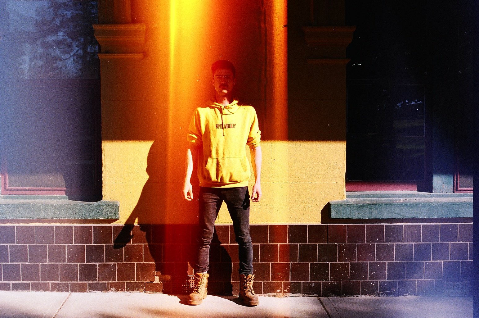 Young man standing against a yellow and brown wall at night, casting a shadow, wearing a yellow hoodie with 'KNOWBODY' written on it, black jeans, and tan boots.