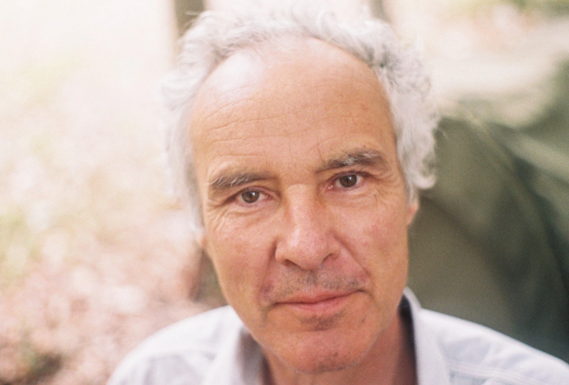 Close-up of a middle-aged man with gray hair, tan skin, and a slight smile, outdoors with blurred natural background.