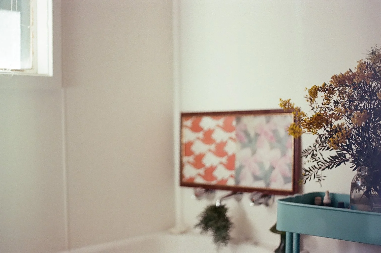 A decorated wall with a framed floral print, a small plant, and a vase filled with yellow flowers on a console table next to a wall and a small window.