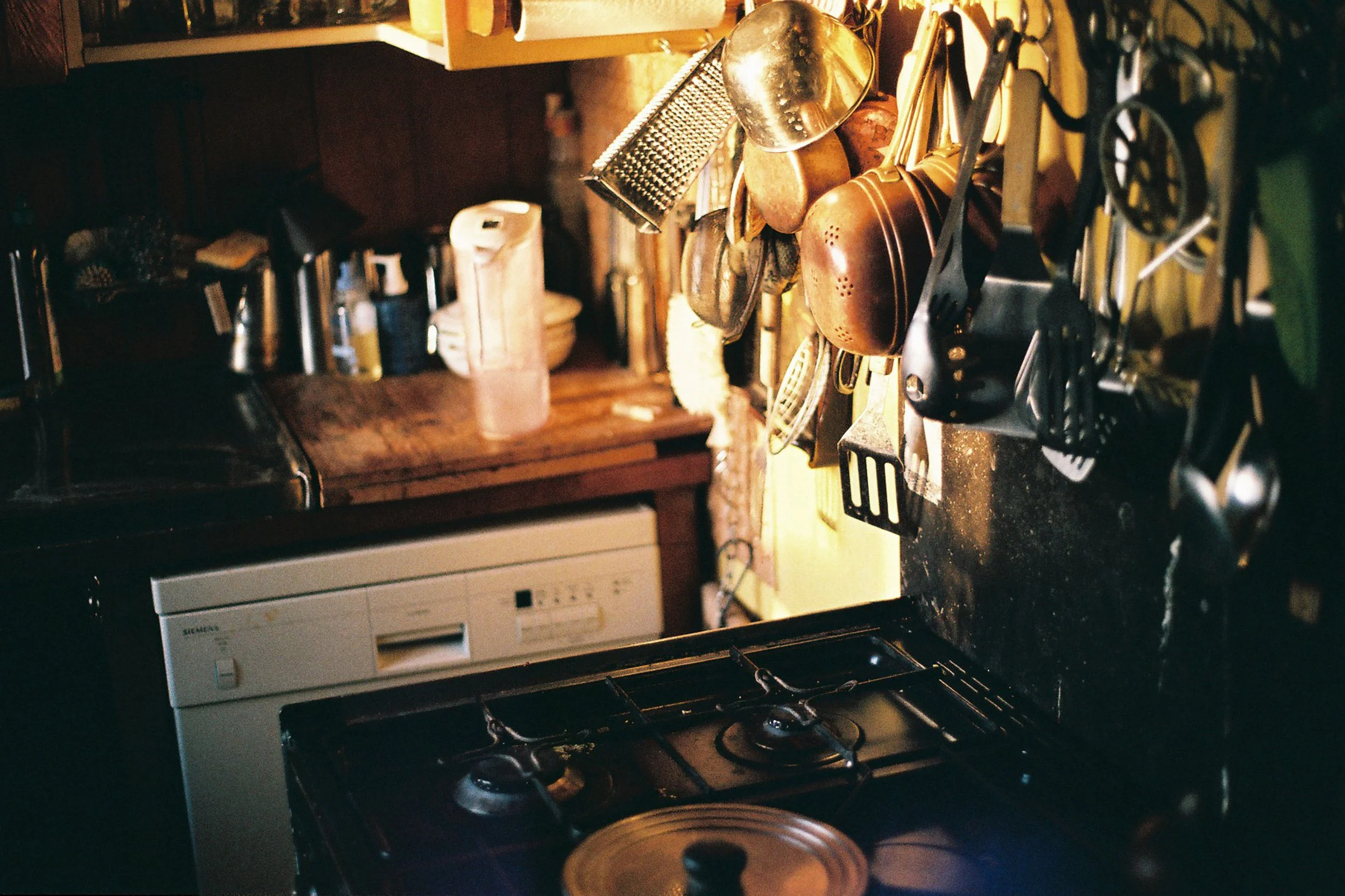 Kitchen scene showing hanging pots, pans, and utensils above a stove and counter with a dishwasher below.
