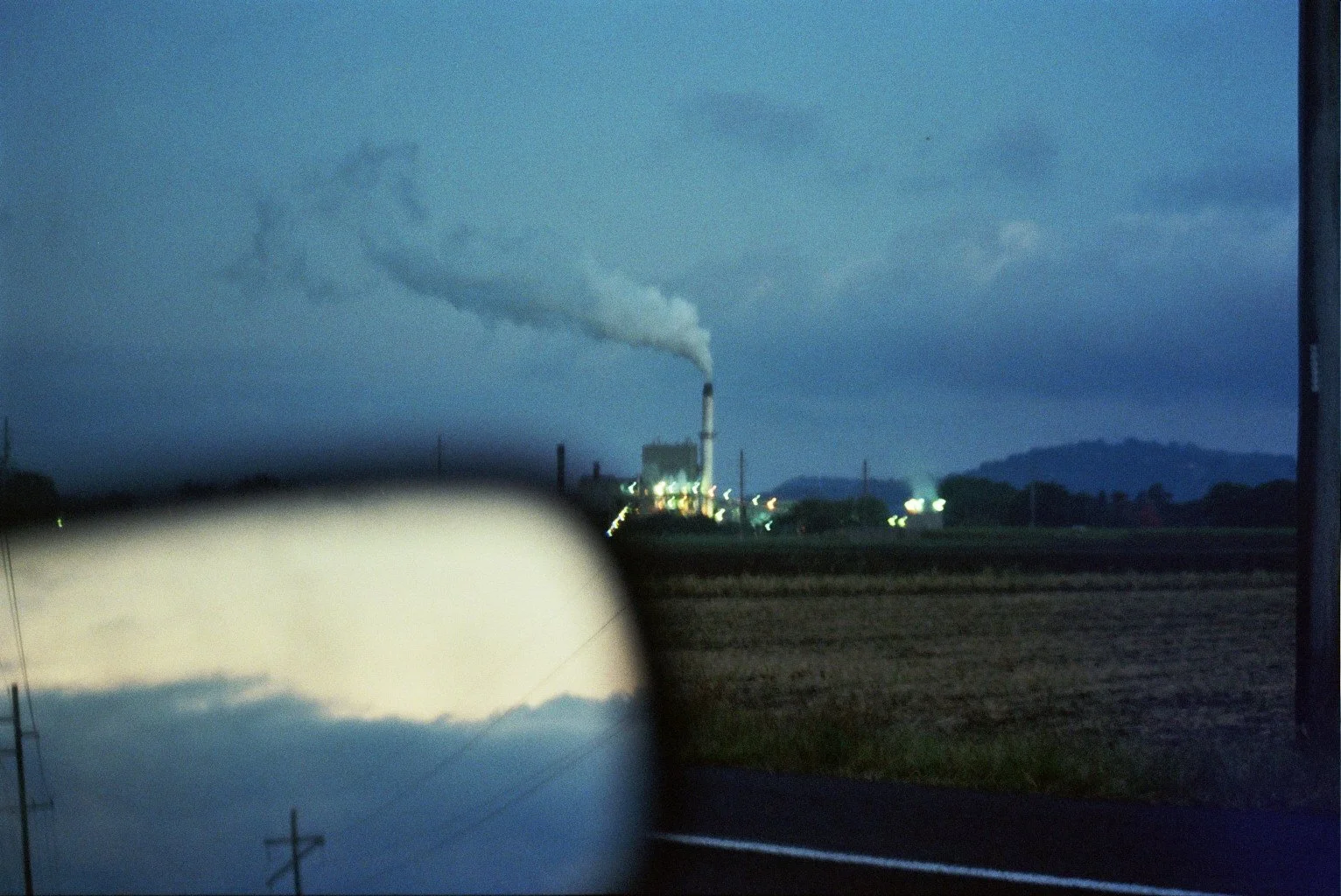 A view of an industrial plant with a smokestack emitting white smoke during dusk or dawn, seen through a car's side mirror and window, with a dark sky and distant hills in the background.