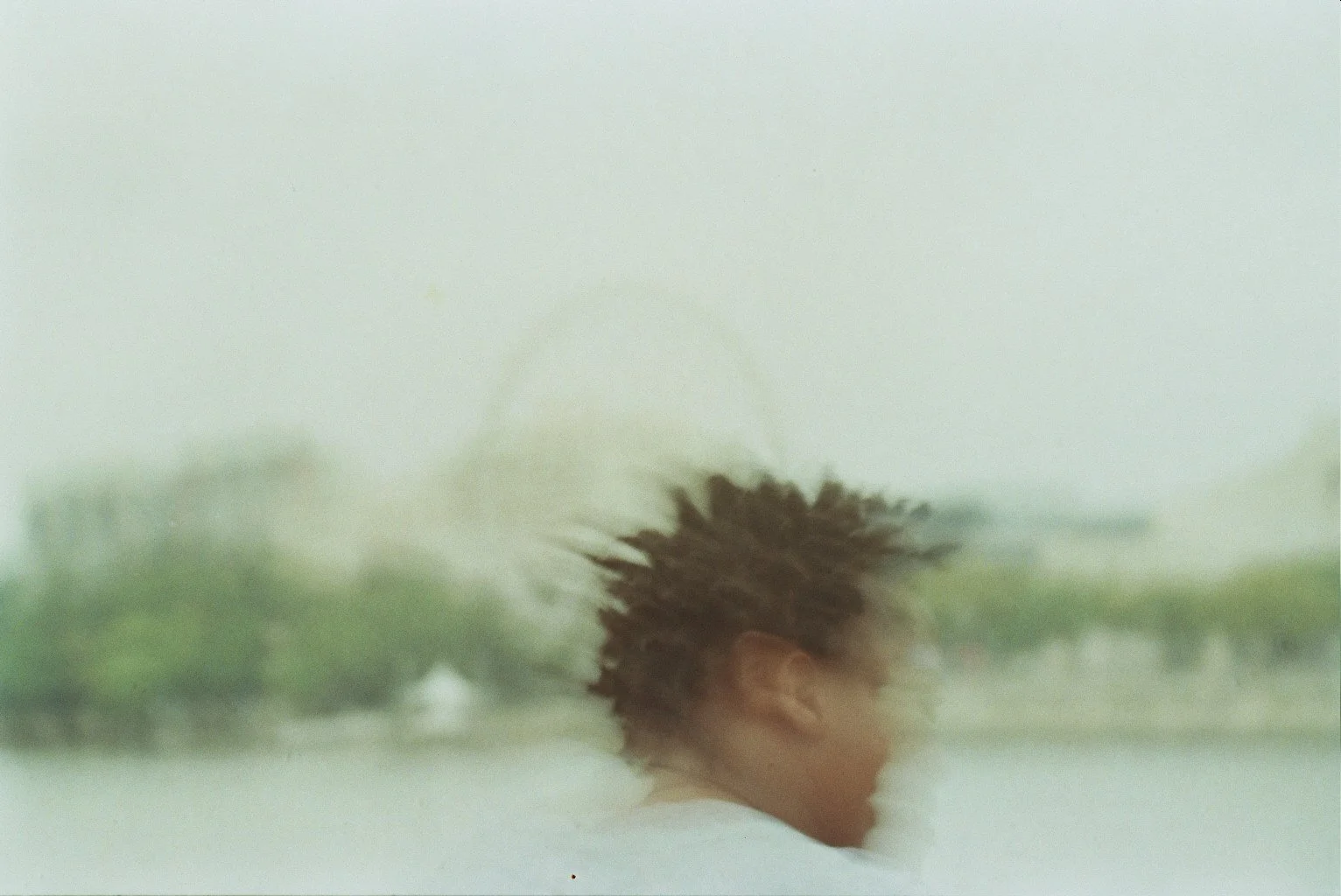 A person with spiky hair standing outside in the rain, blurry background with rain and greenery.