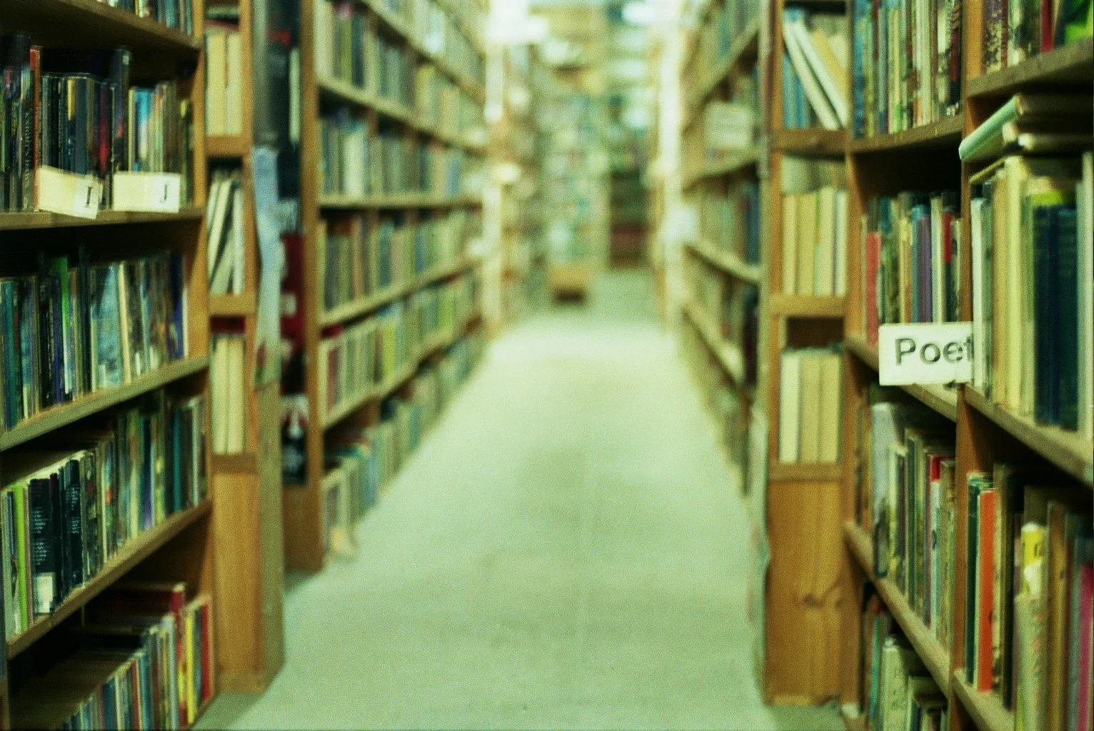 Blurred view of a hallway in a library with wooden bookshelves on either side filled with books; a sign on the right reads 'Poet'.