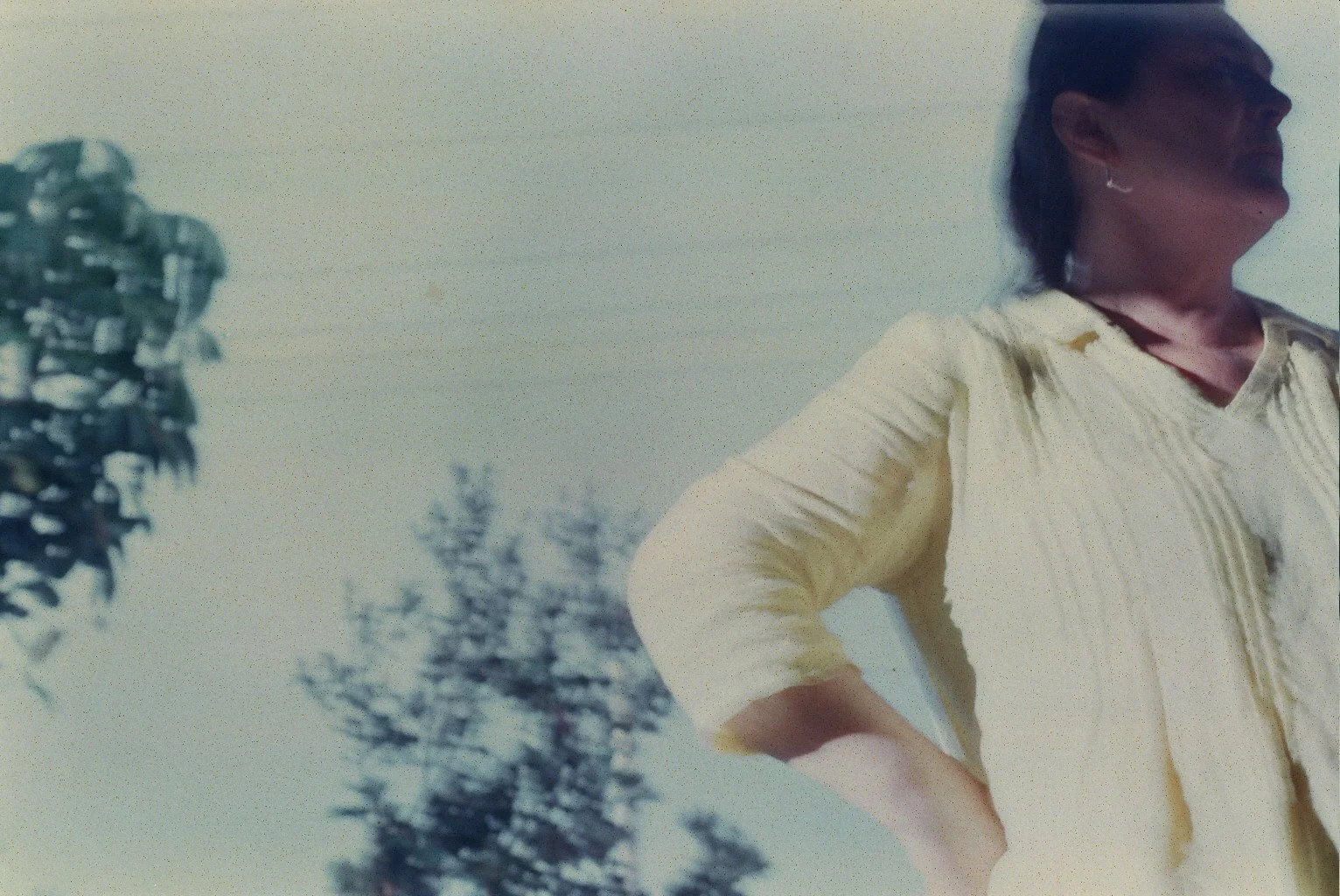 A woman with dark hair tied back, wearing a light yellow shirt, standing outdoors with her hand on her hip, and trees in the background.