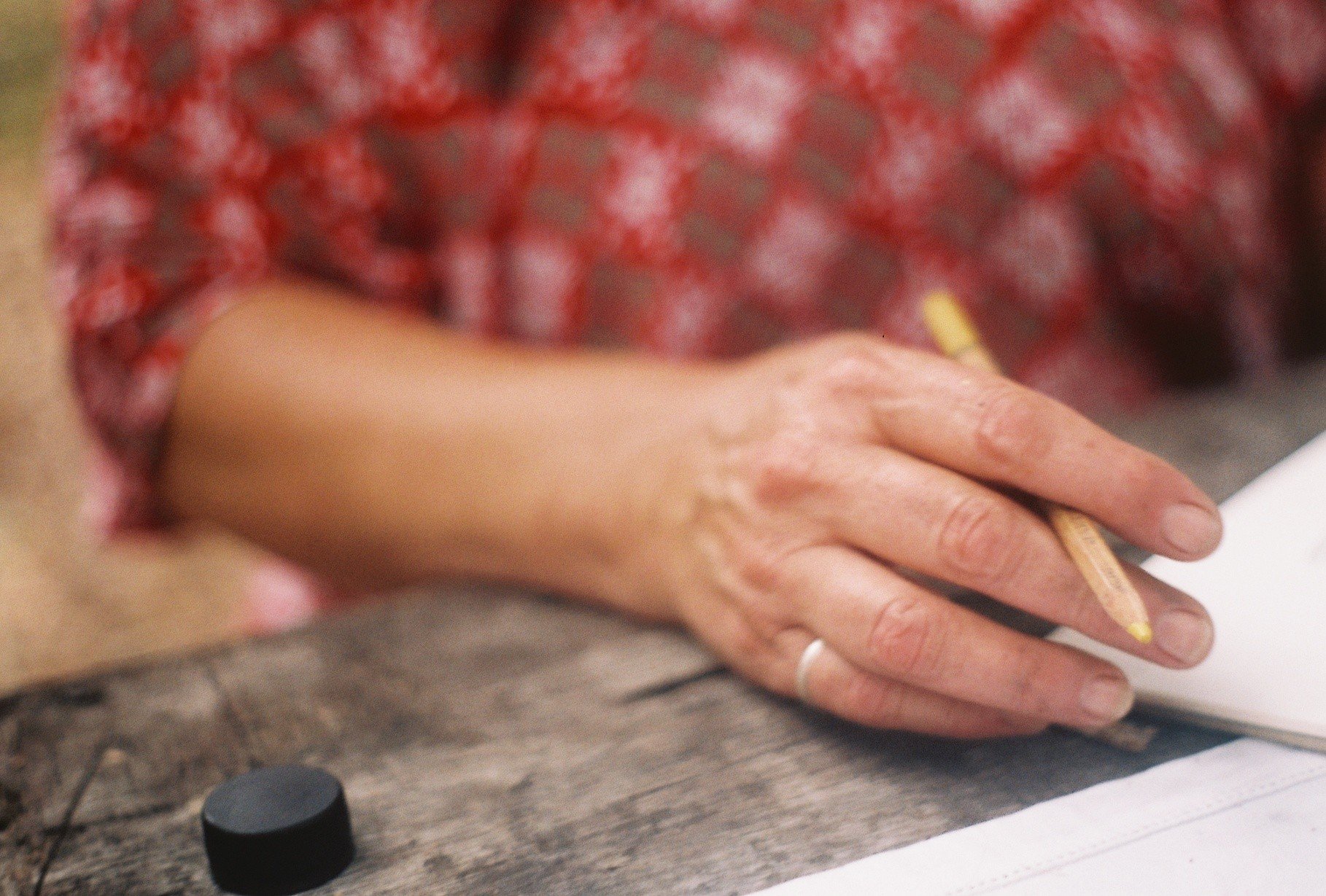 A person with a painted yellow fingernail holds a wooden stick, resting their hand on a wooden surface with a black bottle cap nearby. The person wears a red and blue patterned shirt.