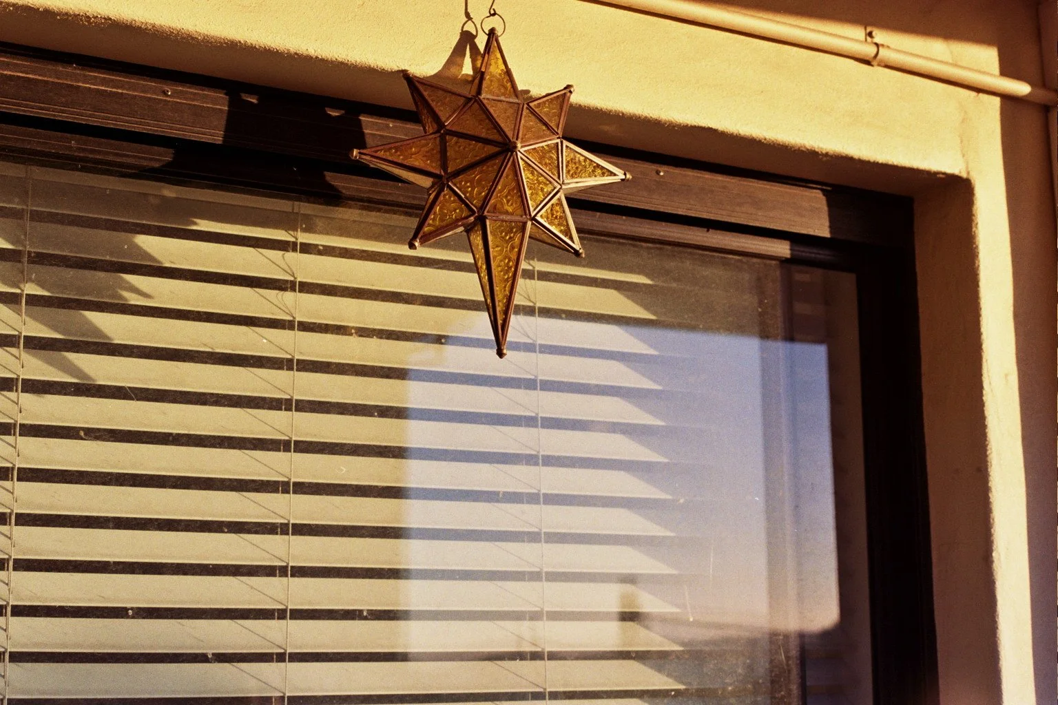A star-shaped hanging lantern with yellow glass panels, attached above a window with horizontal blinds. The window frame is black, and sunlight casts shadows of the blinds onto the glass.