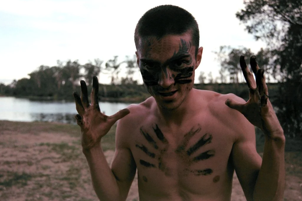 Young man with short hair and black face paint, shirtless, posing outdoors near a body of water in the evening or early morning.