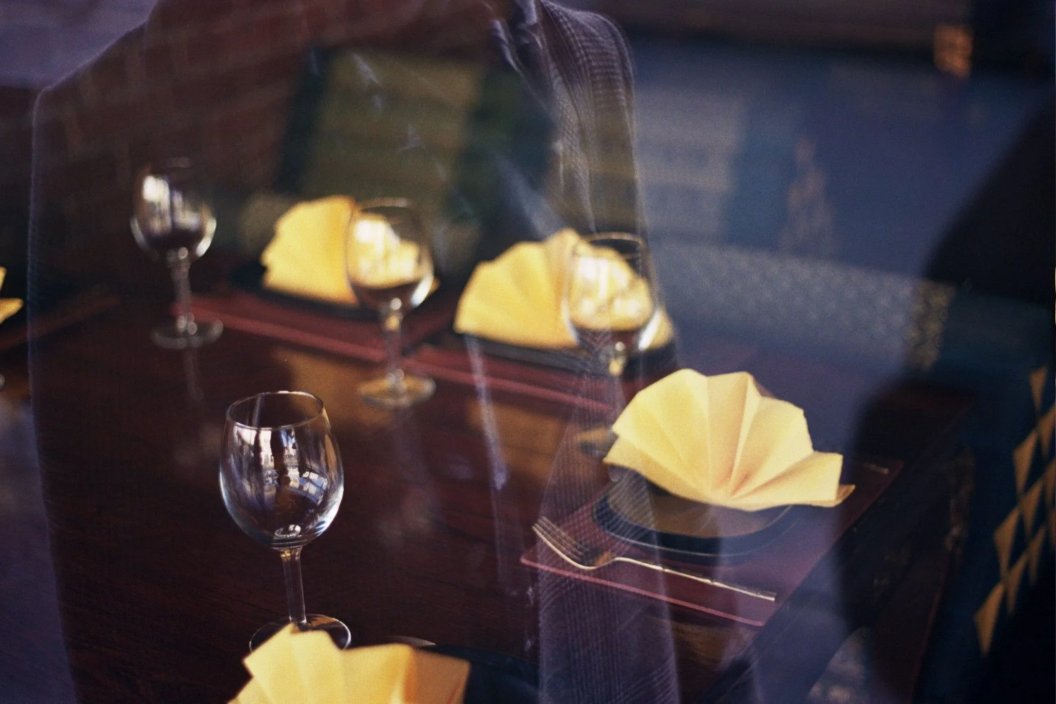 A restaurant table set with empty wine glasses, yellow folded napkins, and silverware, viewed through a window with reflections.