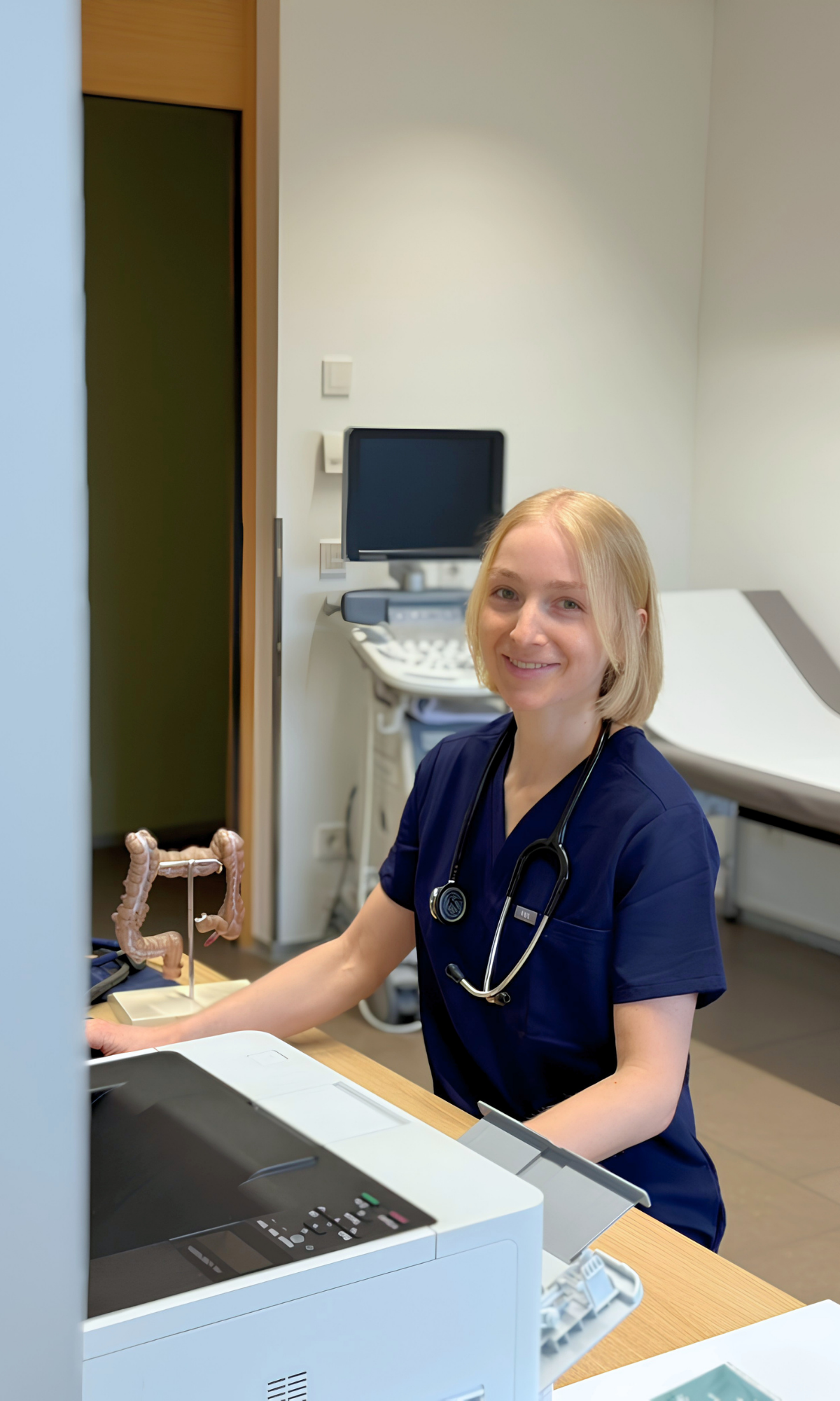 Dr. Julia Längle, Ärztin für evidenzbasierte Allgemeinmedizin in Koblach, sitzt lächelnd mit Stethoskop an ihrem Schreibtisch in der Praxis im Hintergrund ein Modell eines Darms sowie ein Ultraschallgerät.