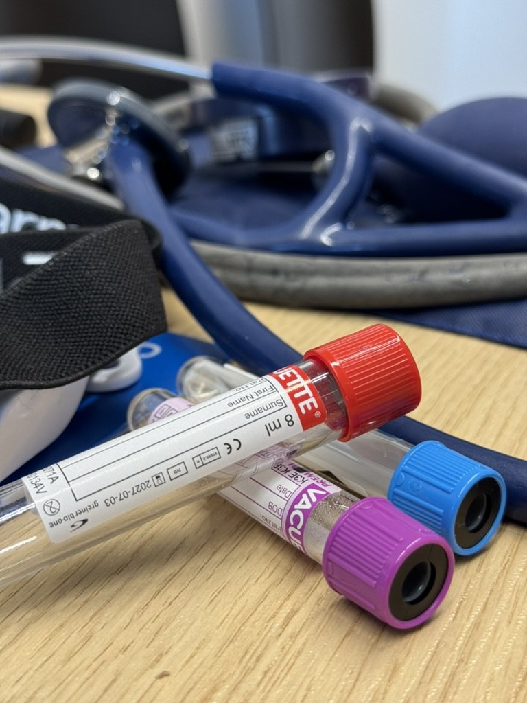 Blutprobenröhrchen mit farbigen Verschlüssen neben Stethoskop und Blutdruckmanschette – Symbol für Stoffwechselanalyse und Laborwerte.
