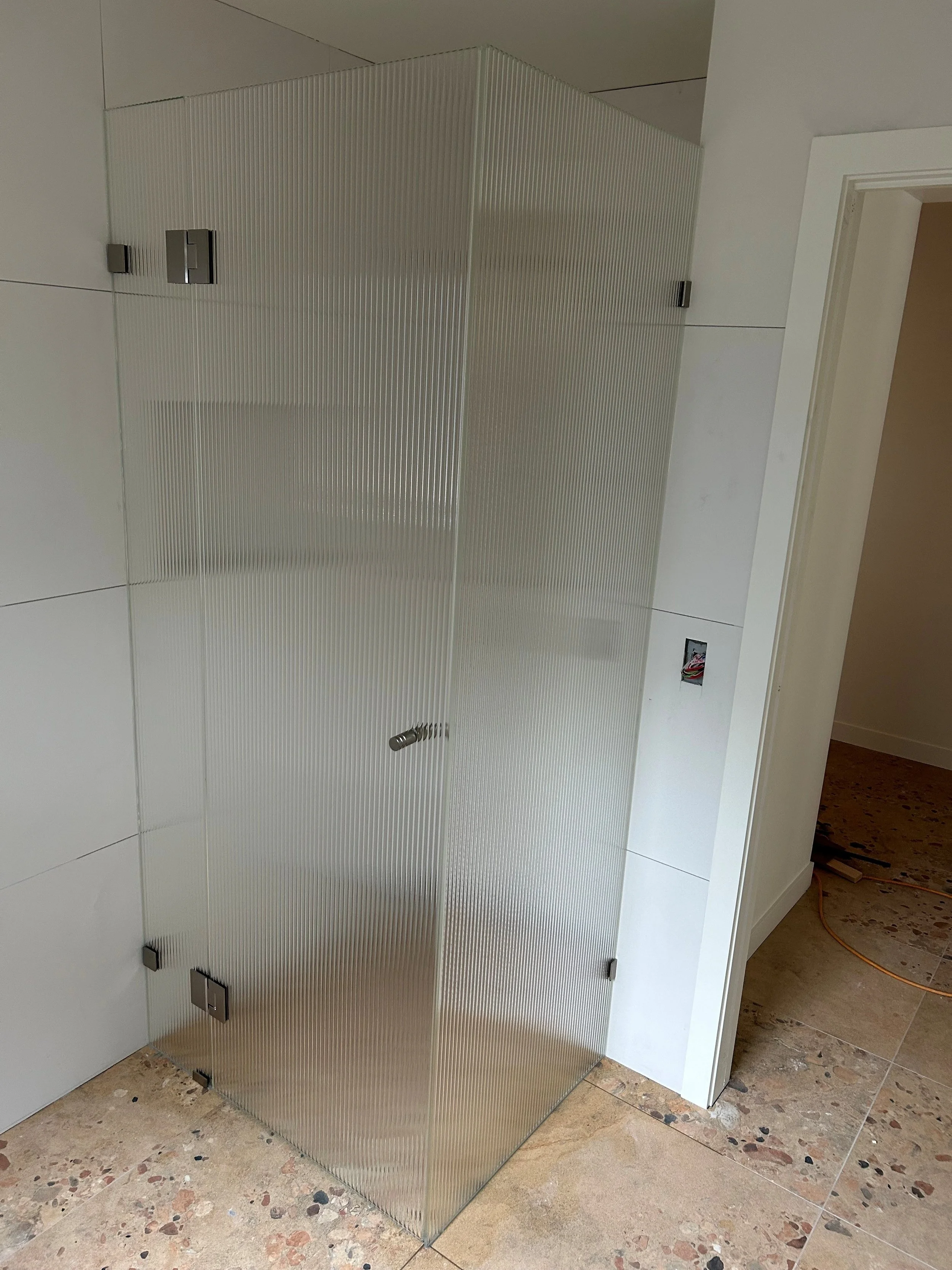 Framesless  shower screen with frosted glass panels and metal hinges on a tiled bathroom floor.