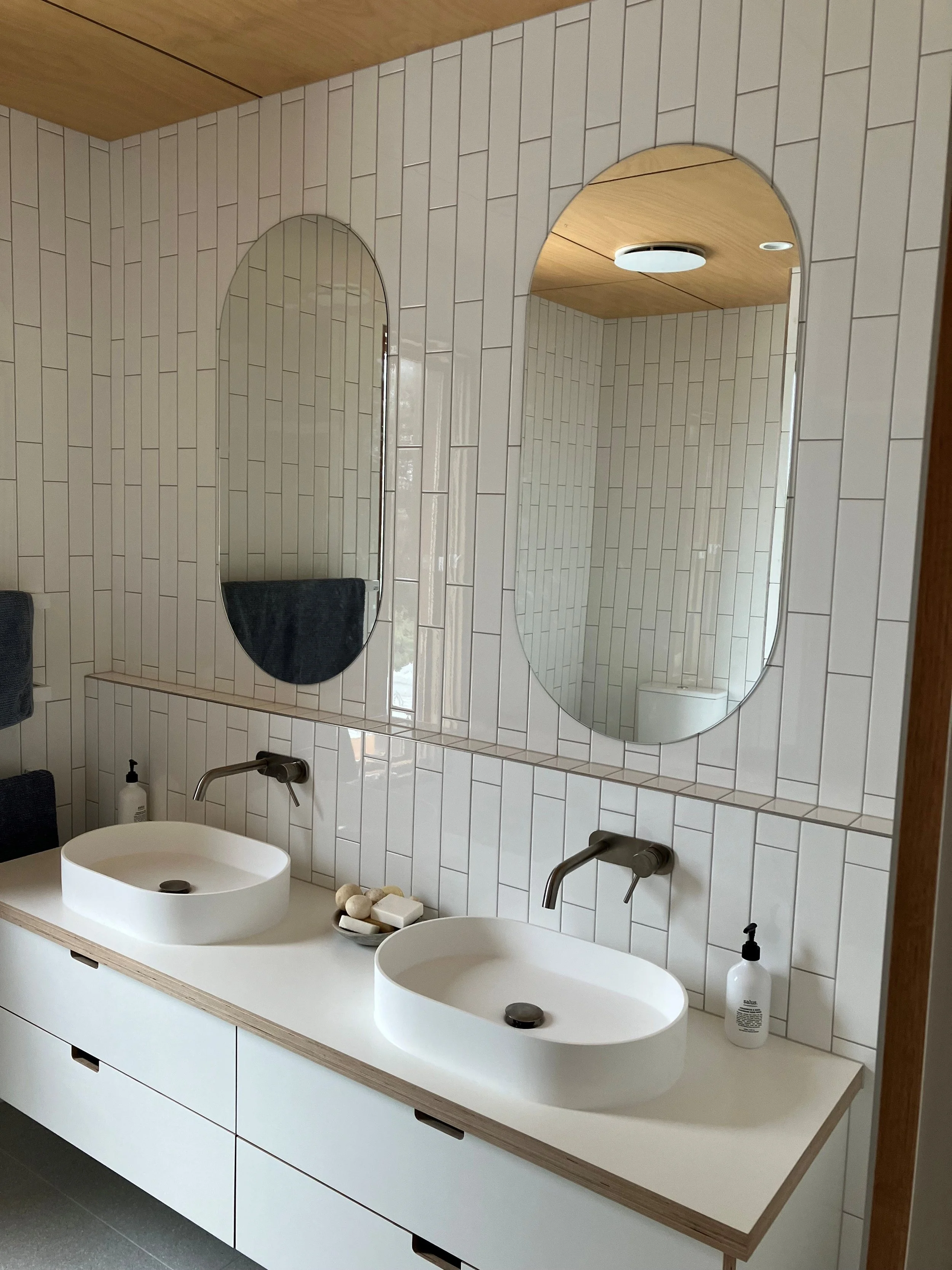 Modern bathroom with two white oval sinks on a light wood vanity, large mirrors on a beige tiled wall, and a wooden ceiling with a circular light fixture.