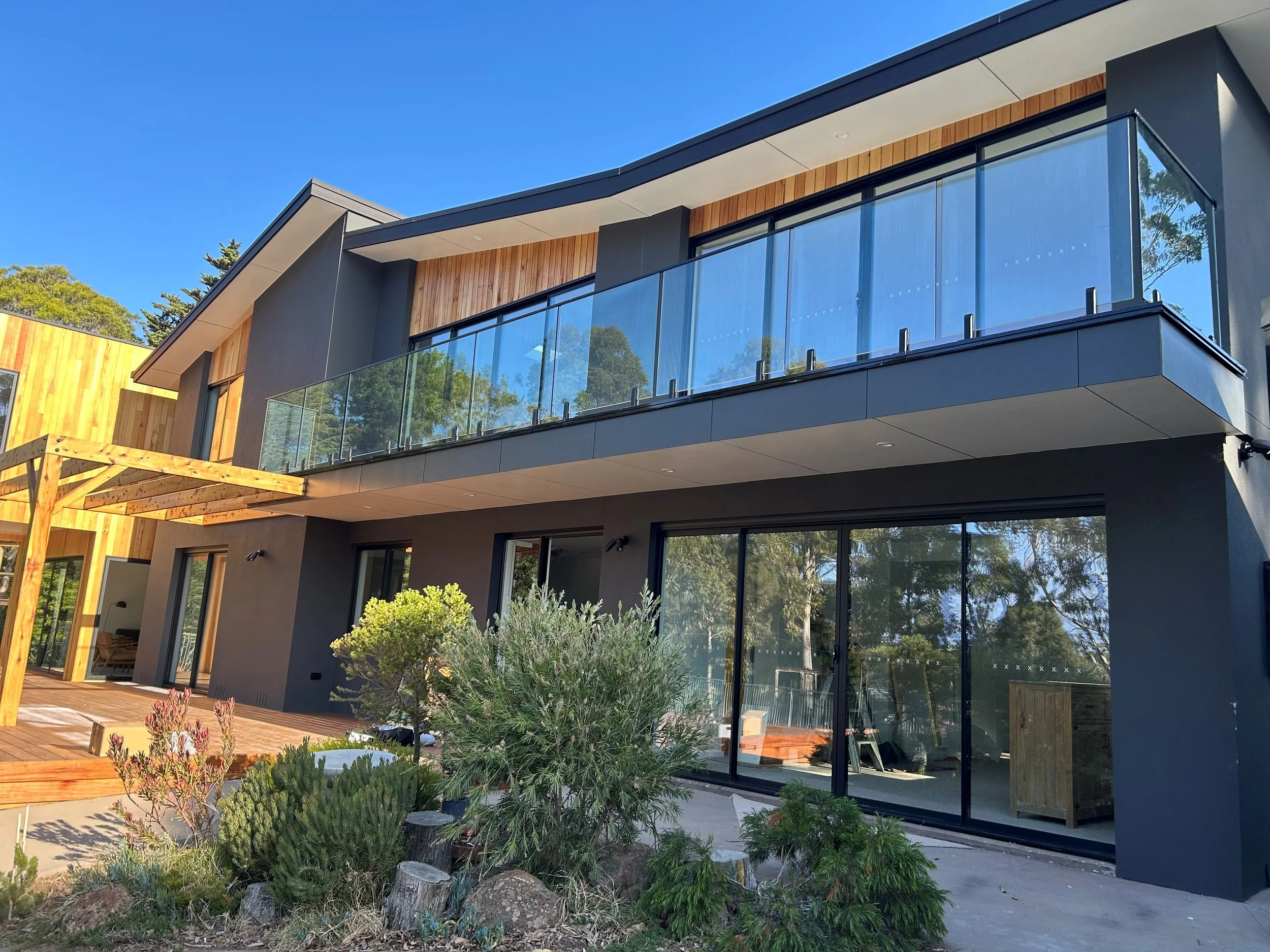 Modern house with large glass balcony, wooden accents, and landscaped garden with bushes and trees in the foreground.