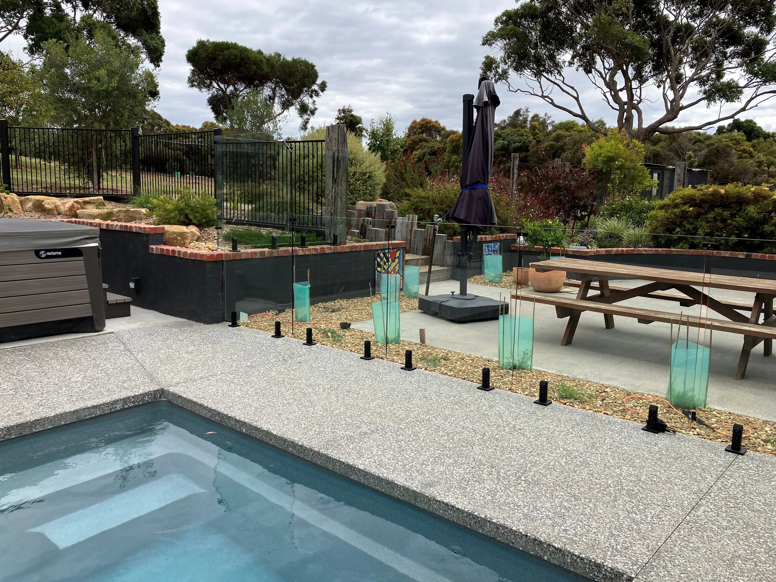 An outdoor backyard scene with a swimming pool in the foreground, a wooden picnic table, a patio umbrella, and a landscaped garden with trees and bushes behind a black fence.
