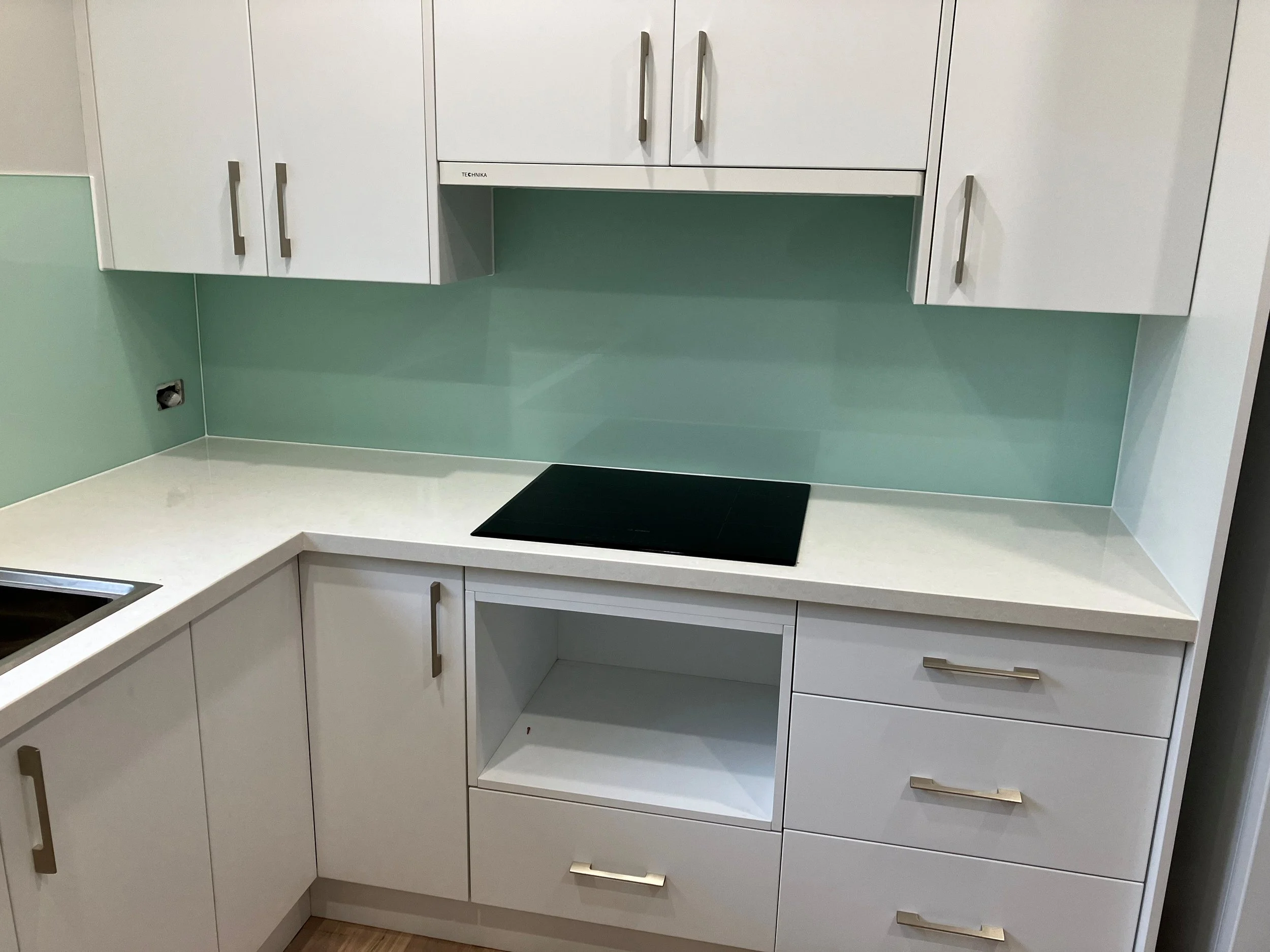 White kitchen cabinets with silver handles and a black cooktop on white countertop against a pale green wall.