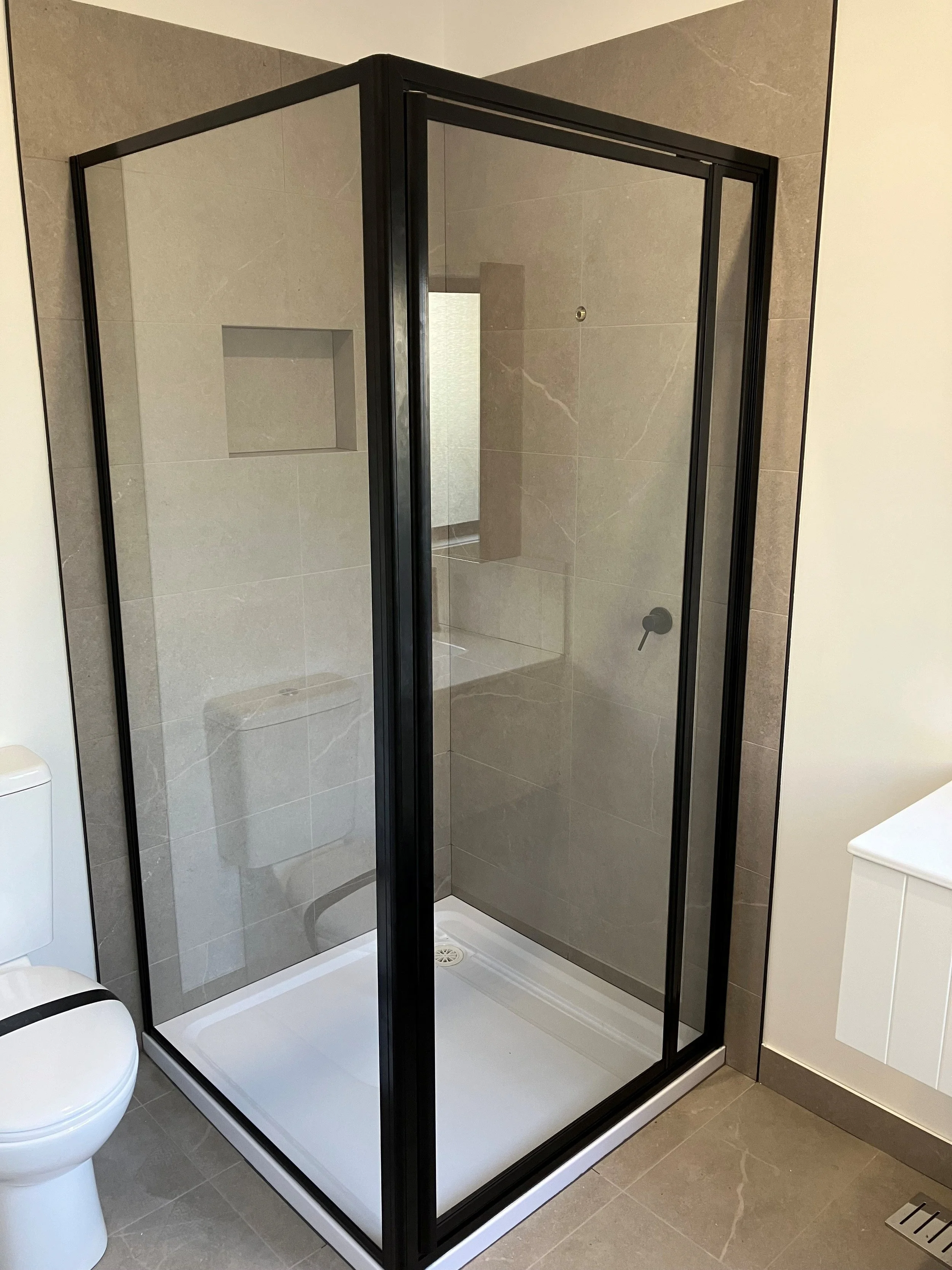 A modern bathroom shower with glass walls and black metal frame, beige tile walls, a built-in niche for toiletries, and a white shower base.