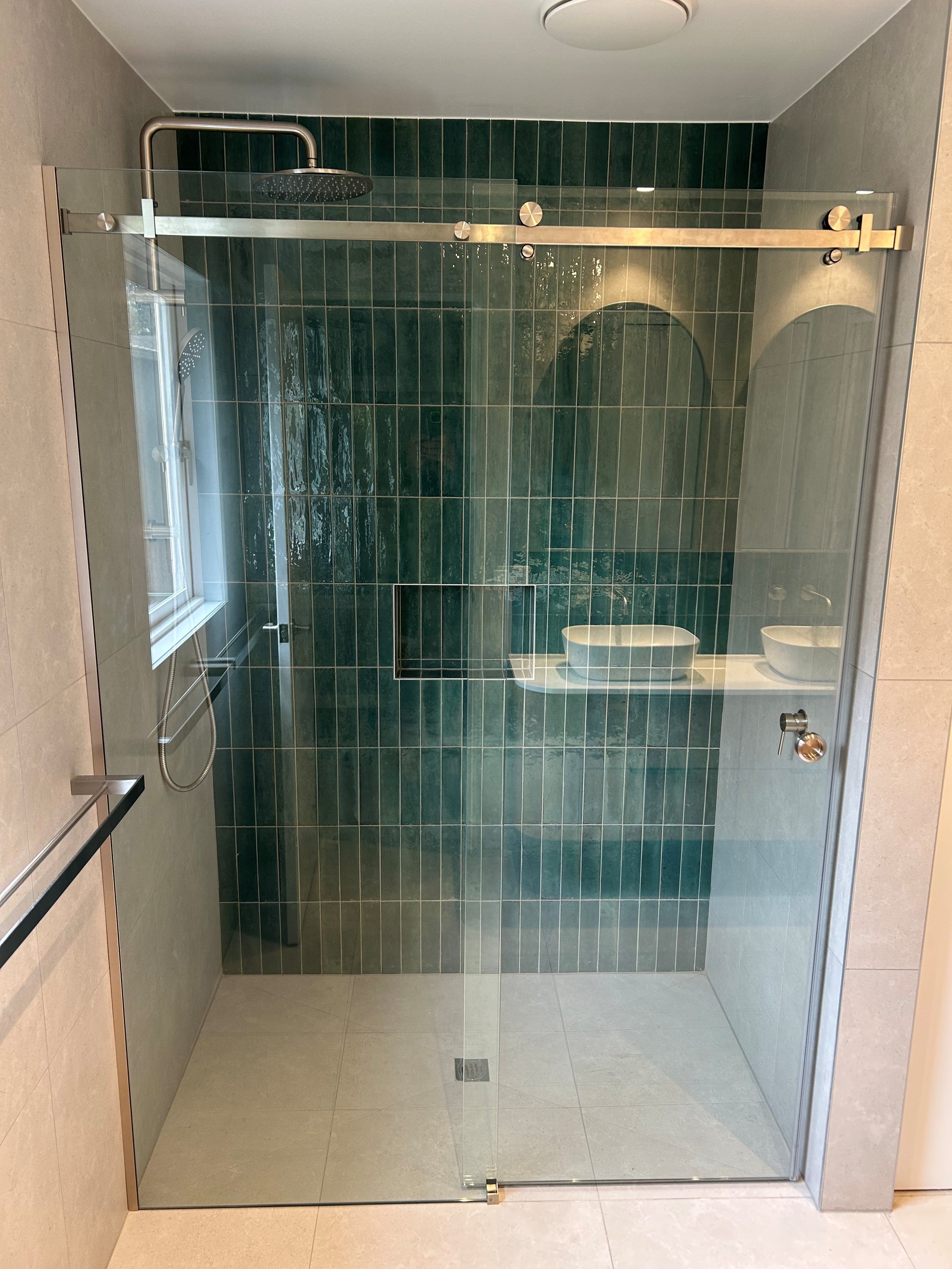 Glass shower enclosure with green tiles on the wall, showerhead, and a handheld shower attachment, located in a modern bathroom.