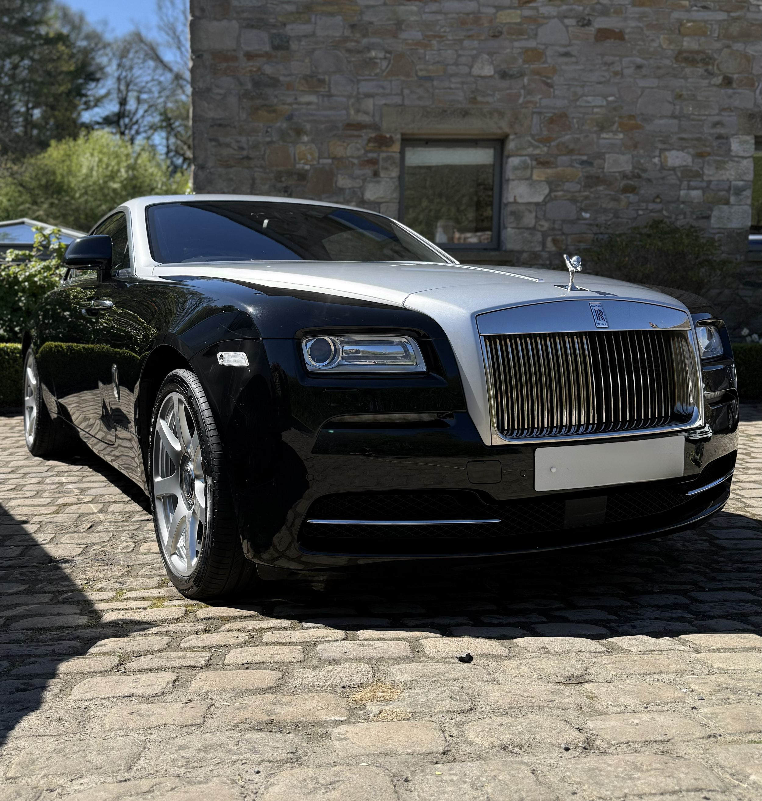 A black and white Rolls Royce luxury car parked on a cobblestone street in front of a stone building with a window. Trees are in the background under a clear sky.