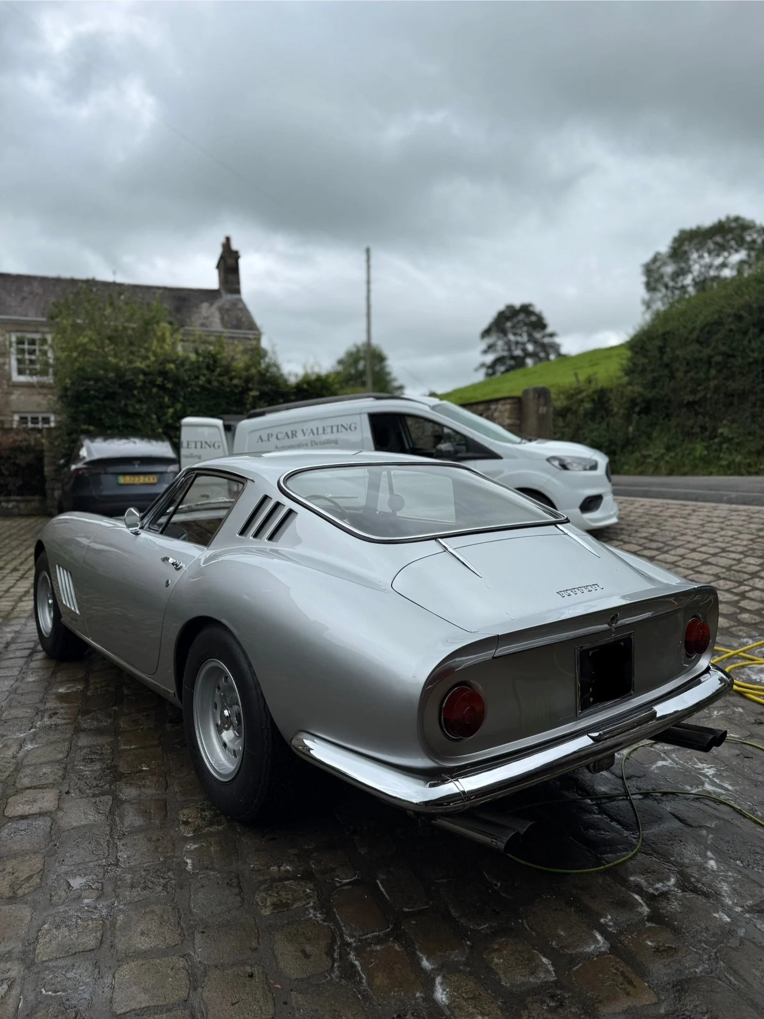 A silver sports car parked on a cobblestone driveway, with a white van and a black car behind it, in a rural setting with grassy hills, trees, and a cloudy sky.