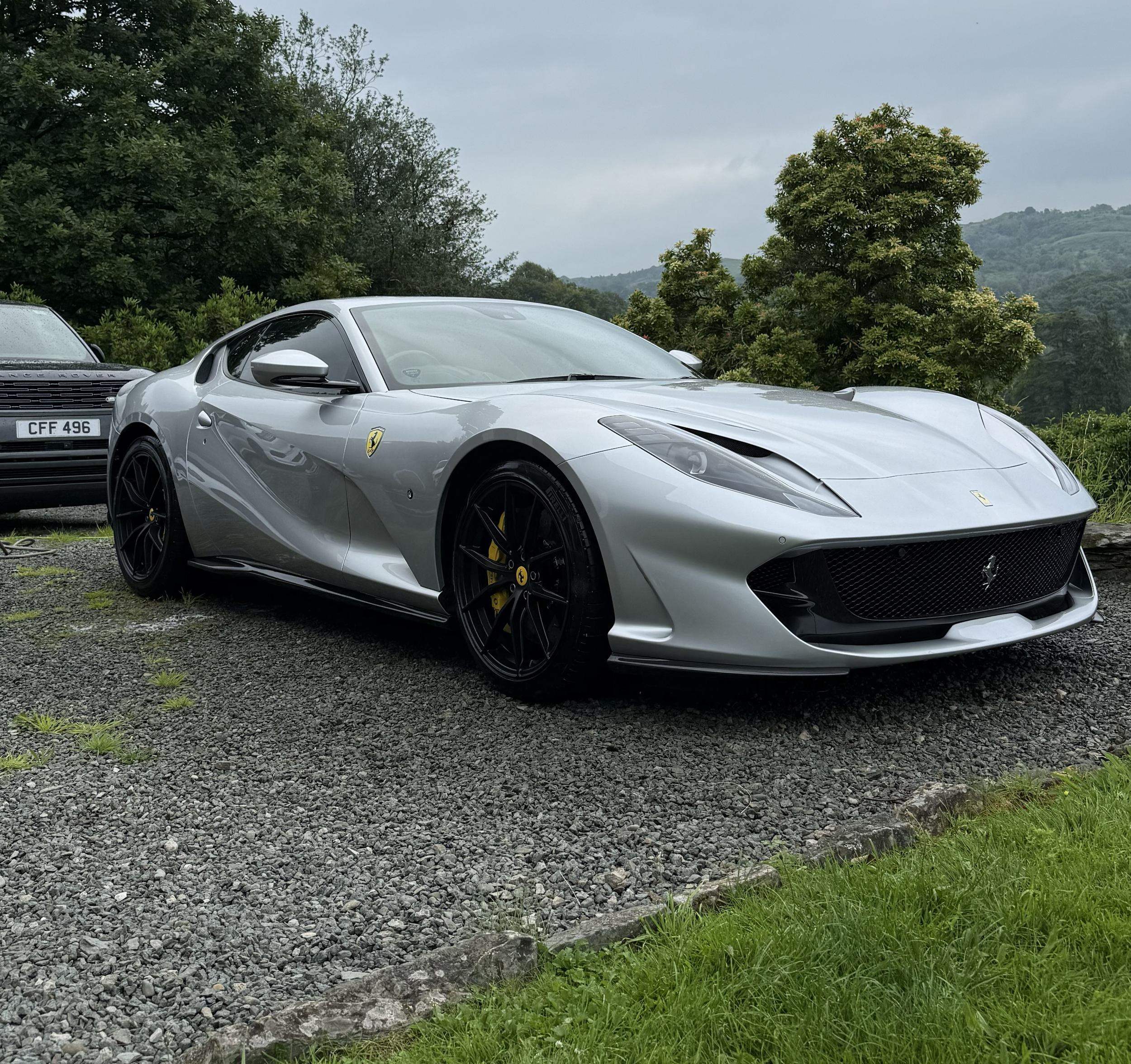 A white Ferrari sports car parked outdoors on gravel next to a grey Land Rover with a white roof. The background features lush green trees and hills under a cloudy sky.
