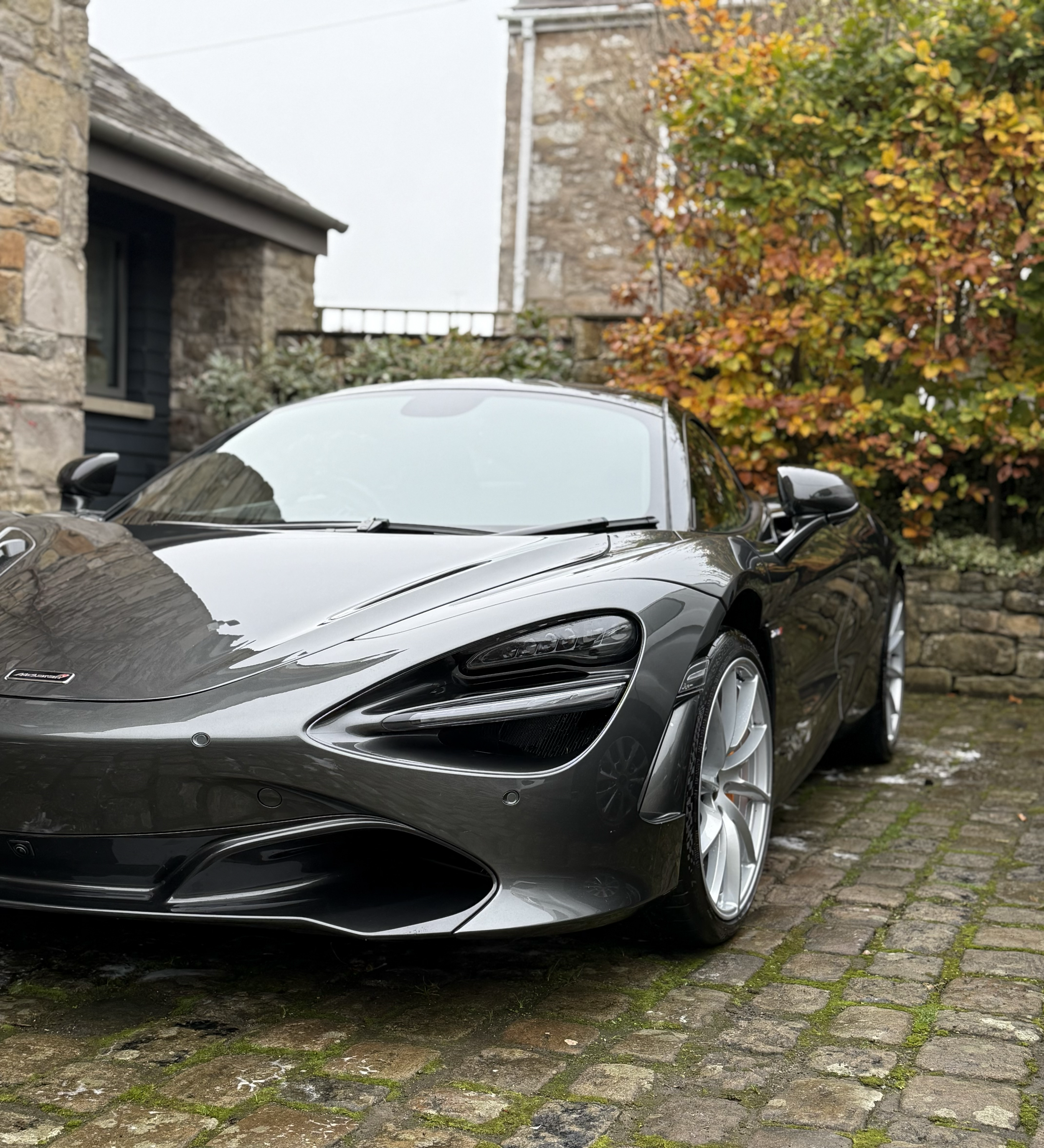A black sports car with a sleek design is parked on a cobblestone driveway. The background features a stone building with a darkly painted window frame, a stone wall, and colorful autumn foliage.