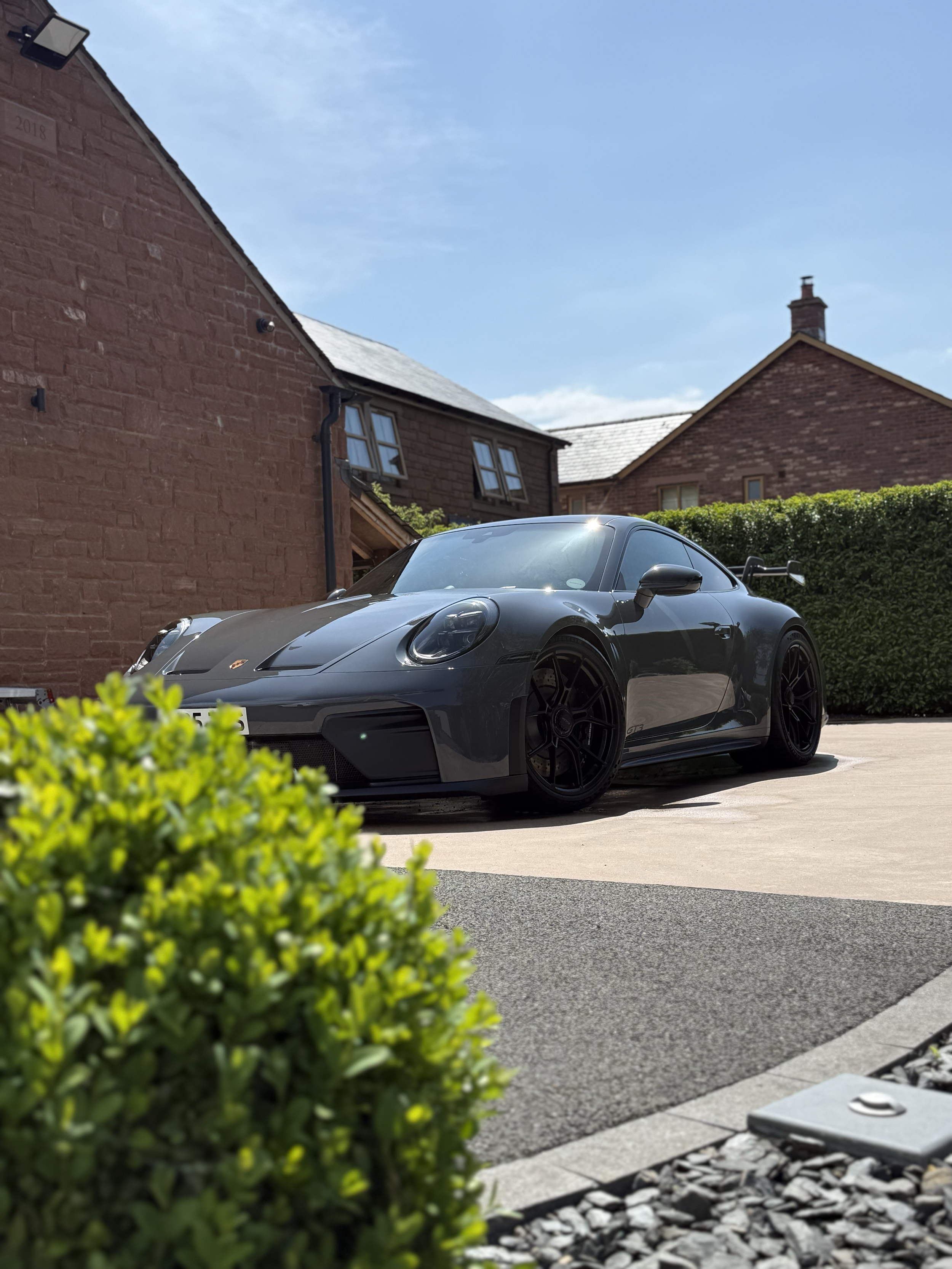 A black Porsche sports car parked in a driveway in front of brick houses with small windows and a hedge; some greenery in the foreground and a blue sky with wispy clouds.