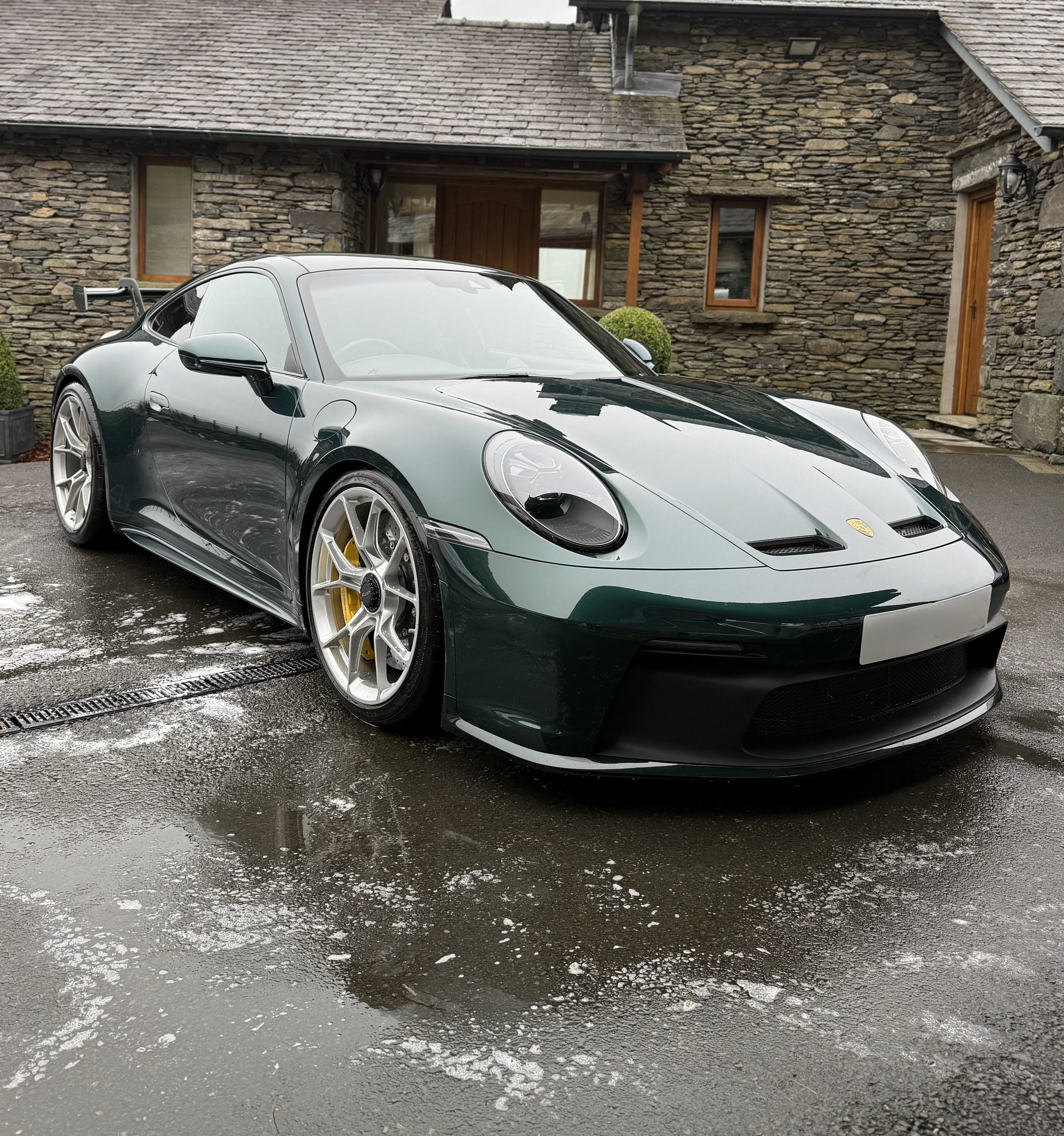 Close-up of a black Porsche sports car parked on a wet driveway with patches of ice, in front of a stone house with wooden window frames and a wooden door.