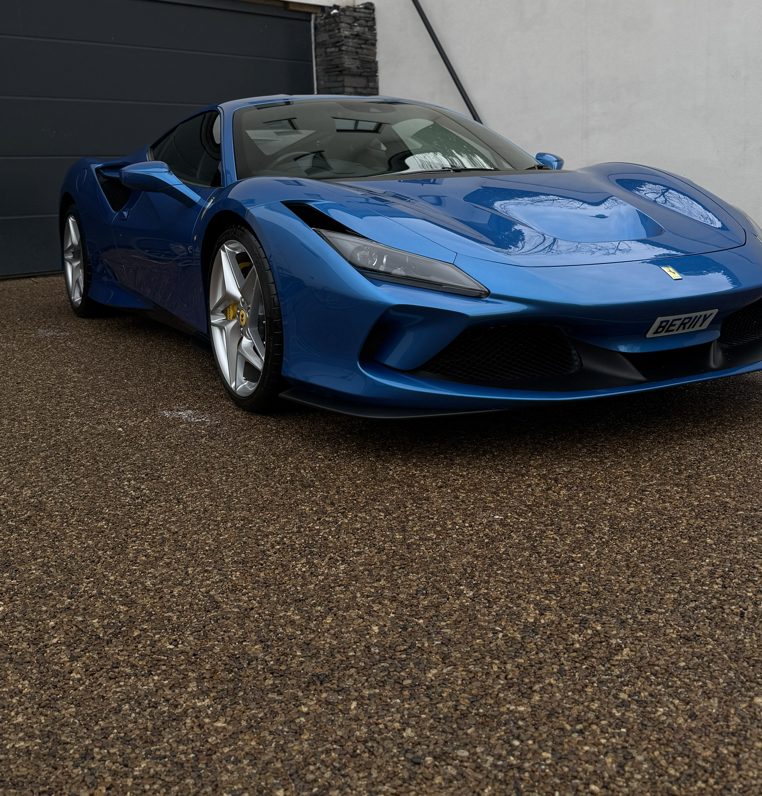 A blue Ferrari sports car parked outside, with a modern dark door and a brick wall in the background.